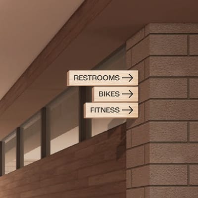 Interior wayfinding signage rendering for Bloc House, showing three stacked directional signs mounted on a warm brick column. Signs read "Restrooms," "Bikes," and "Fitness," each with a right-pointing arrow, rendered in a natural wood finish against the building's dark wood and brick corridor.