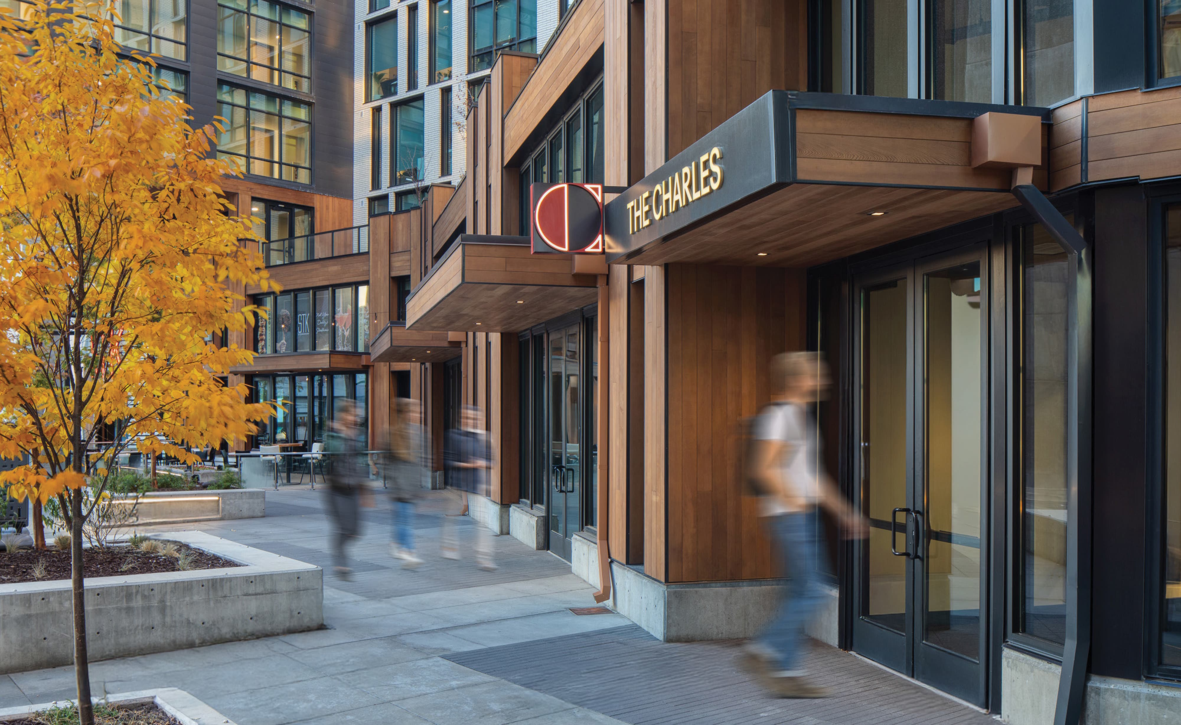 RSM Design exterior entry signage and placemaking for The Charles at The West Quarter in Salt Lake City, featuring illuminated dimensional lettering and a bold geometric logo medallion mounted on a warm wood and steel canopy above a glass entry door, set within an activated pedestrian plaza with motion-blurred figures, autumn foliage, and concrete seating in the foreground, illustrating how environmental graphic design, visual identity, landmark signage, and branded environments create a welcoming, human-centered sense of arrival for mixed-use hospitality, residential community, food and beverage, and urban streetscape markets.