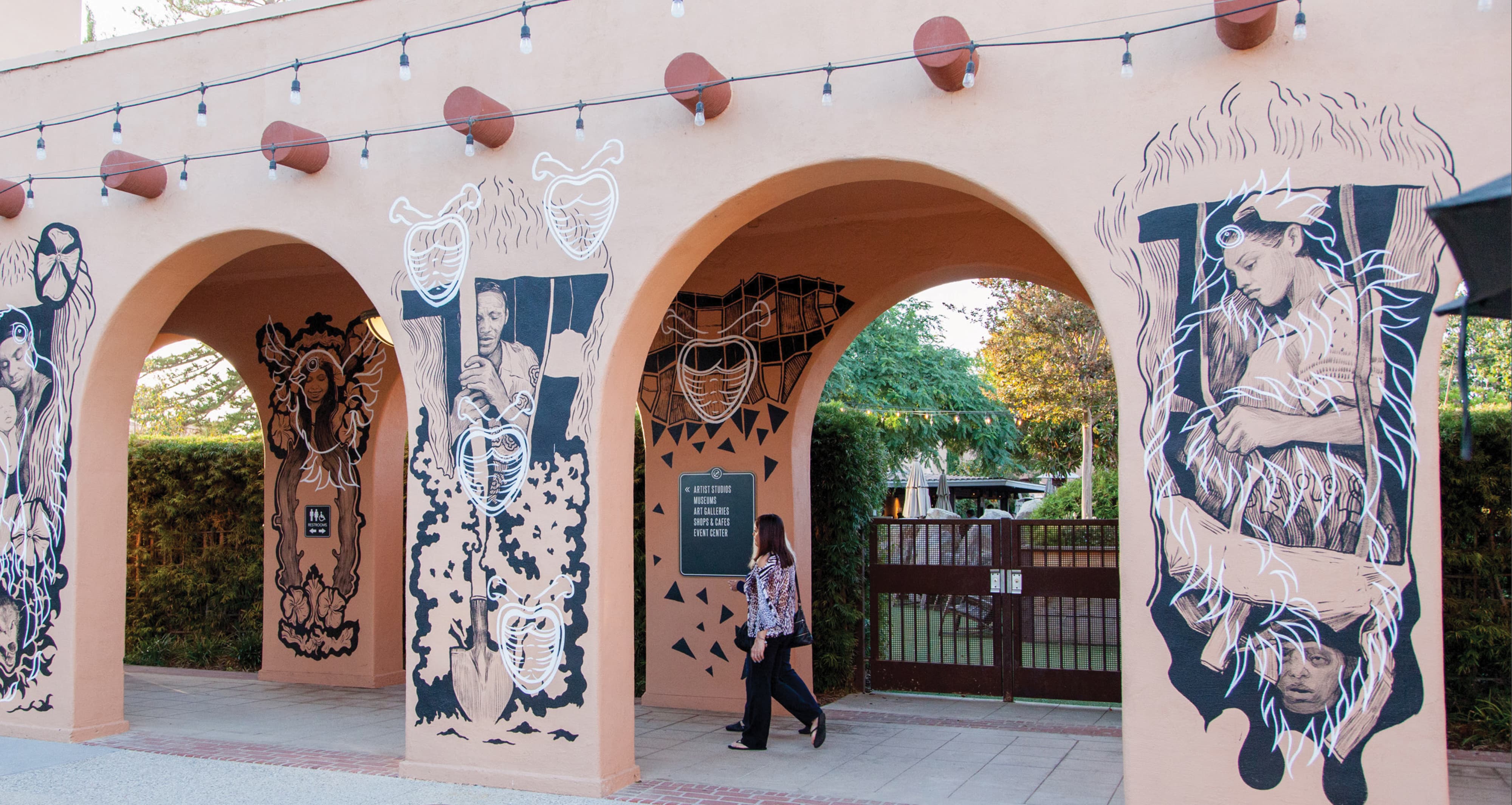Row of pink stucco archways painted with black-and-white illustrated murals of figures and hearts, string lights overhead, a wayfinding sign for Artist Studios, Museums, and Shops & Cafes, as a woman walks toward a metal gate leading to a garden beyond.