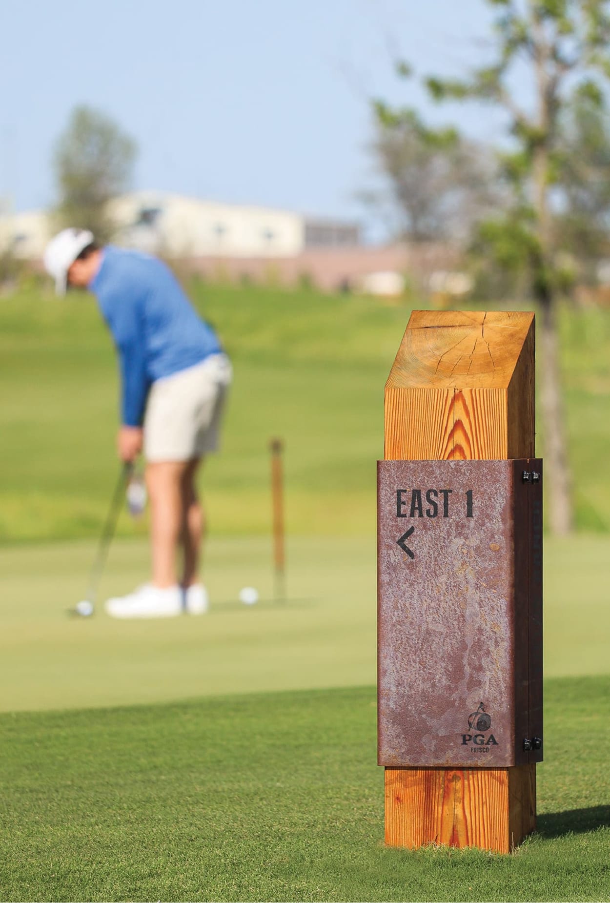 Close-up of PGA Frisco golf course wayfinding marker featuring weathered corten steel panel and wood post with East 1 directional signage, golfer on green in background