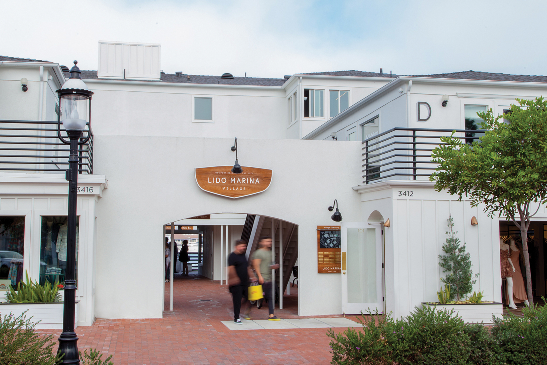 Shoppers walking through Lido Marina Village entrance archway with wood nautical sign, white coastal architecture, brick pathway, and boutique storefronts in Newport Beach, California
