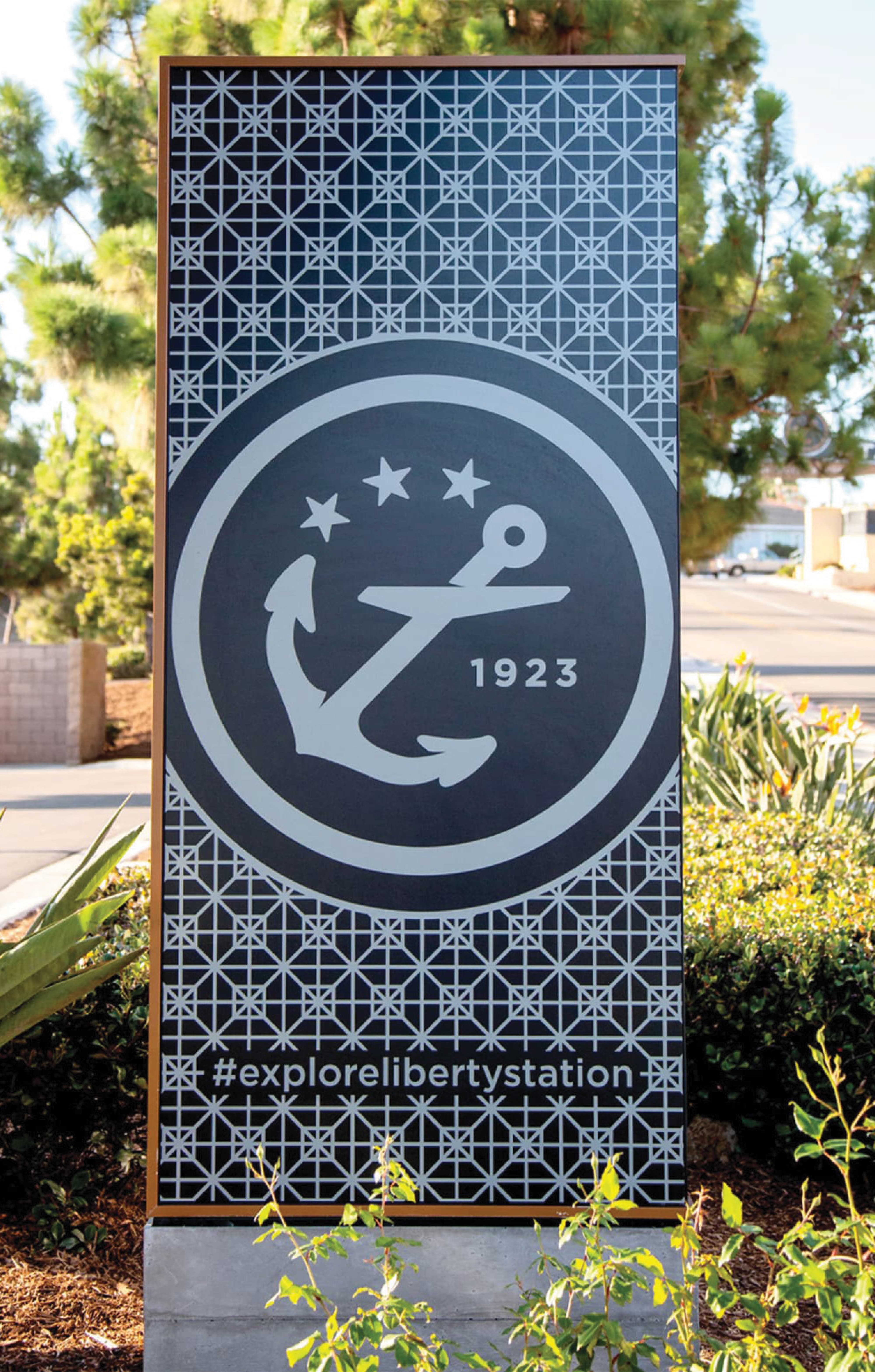 Tall monument sign with a lattice pattern background and a circular anchor emblem with three stars and '1923,' featuring the hashtag '#explorelibertystation' below, set among landscaping with agave and shrubs.