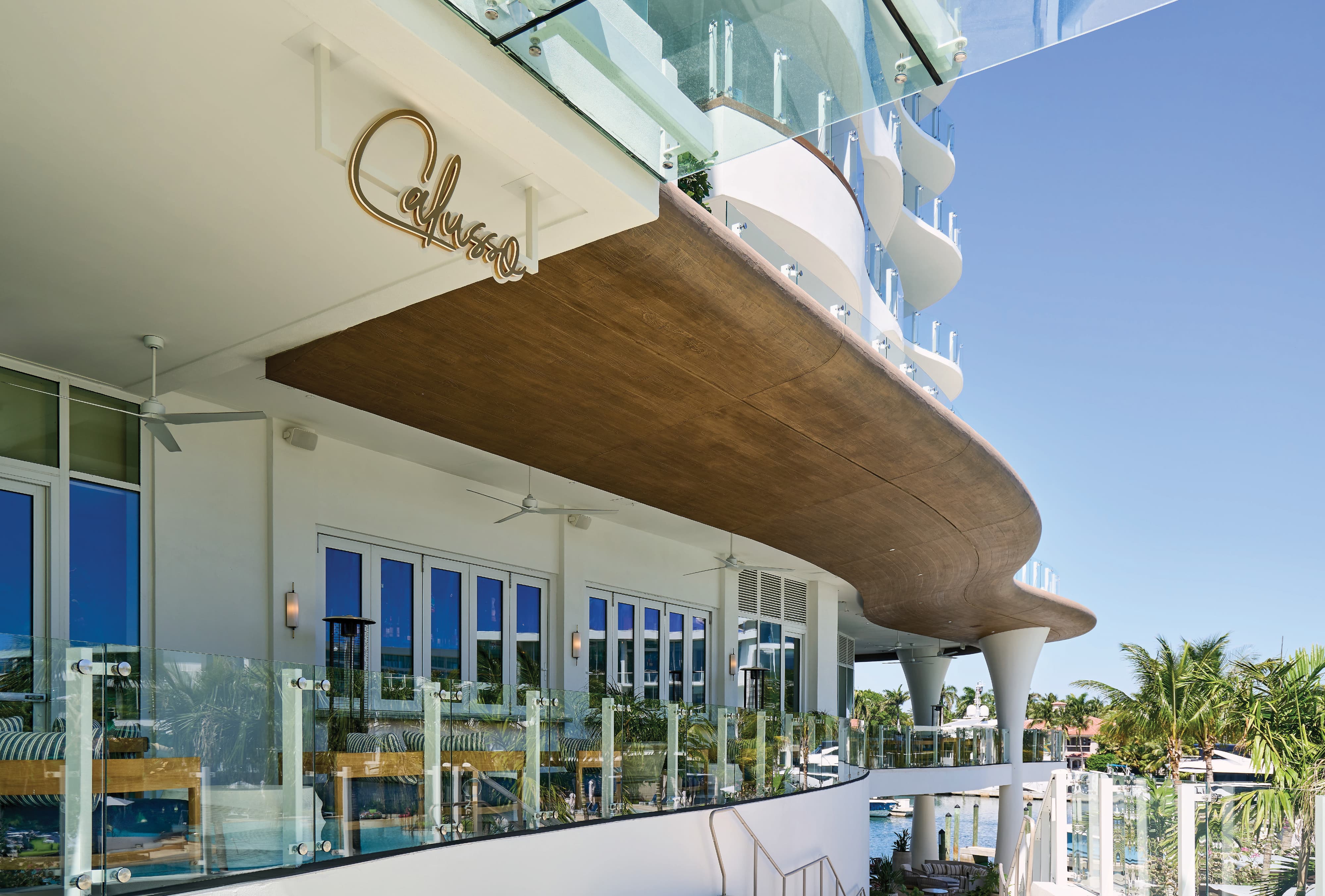 Gold dimensional script logo sign for Calyso restaurant mounted on the exterior facade of a curved cantilevered terrace overlooking the marina and pool at Pier Sixty-Six luxury waterfront resort in Fort Lauderdale, Florida — RSM Design branding and visual identity, wayfinding and signage design, specialty graphics, and branded environments for hospitality, food and beverage, and waterfront destinations.