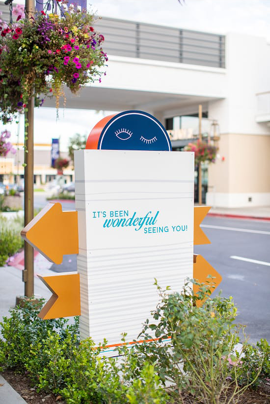 Exterior vehicular directional wayfinding sign at 2nd & PCH in Long Beach, California, featuring a playful illustrated face with winking eyes on a navy semicircle topper, orange arrow blades, and a farewell message reading 'It's Been Wonderful Seeing You!' — an example of RSM Design's placemaking, experiential graphics, and wayfinding signage design that creates moments of delight within a coastal waterfront retail and mixed-use destination.