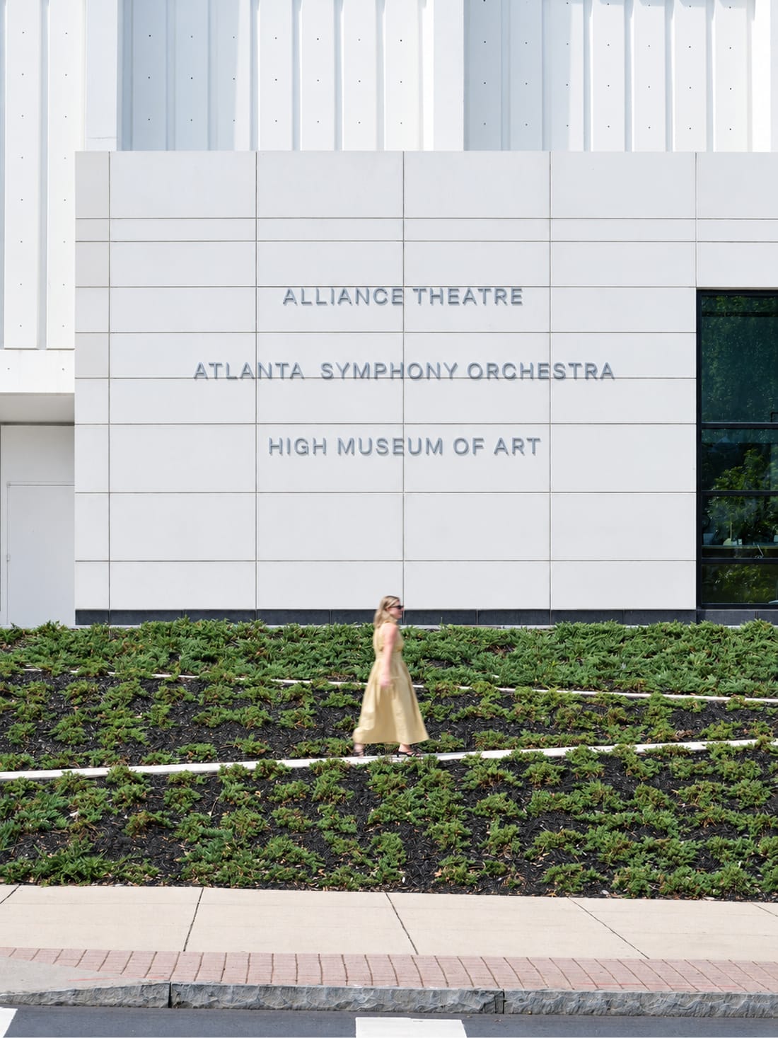 Wall-mounted directory sign listing Alliance Theatre, Atlanta Symphony Orchestra, and High Museum of Art on a white facade above a landscaped groundcover bed at the Woodruff Arts Center – campus wayfinding and environmental graphic design by RSM Design.