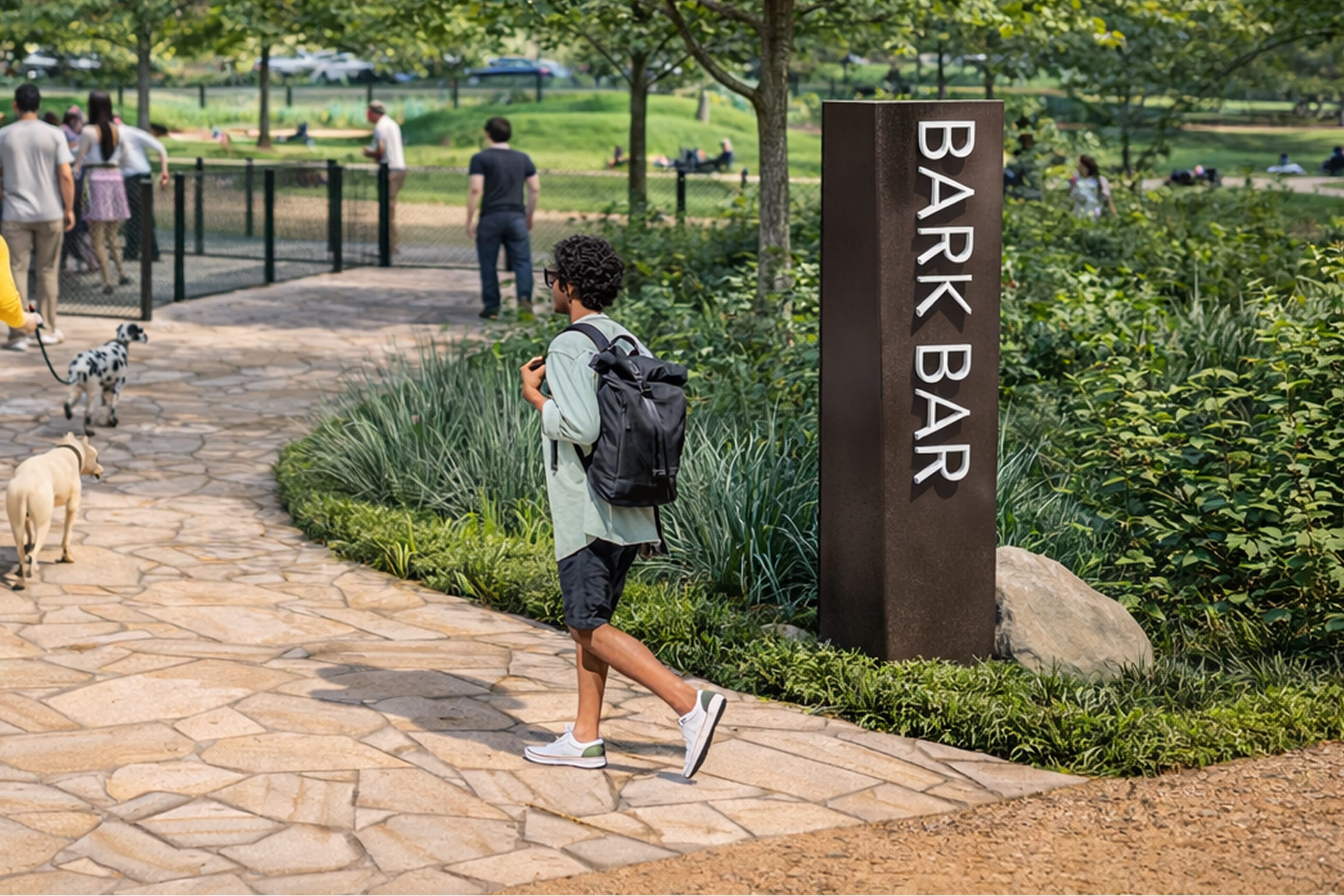Dark monolithic "Bark Bar" identification sign on a stone plaza near a fenced dog park and open-air pavilion, with visitors and dogs gathered nearby — wayfinding and signage design by RSM Design.