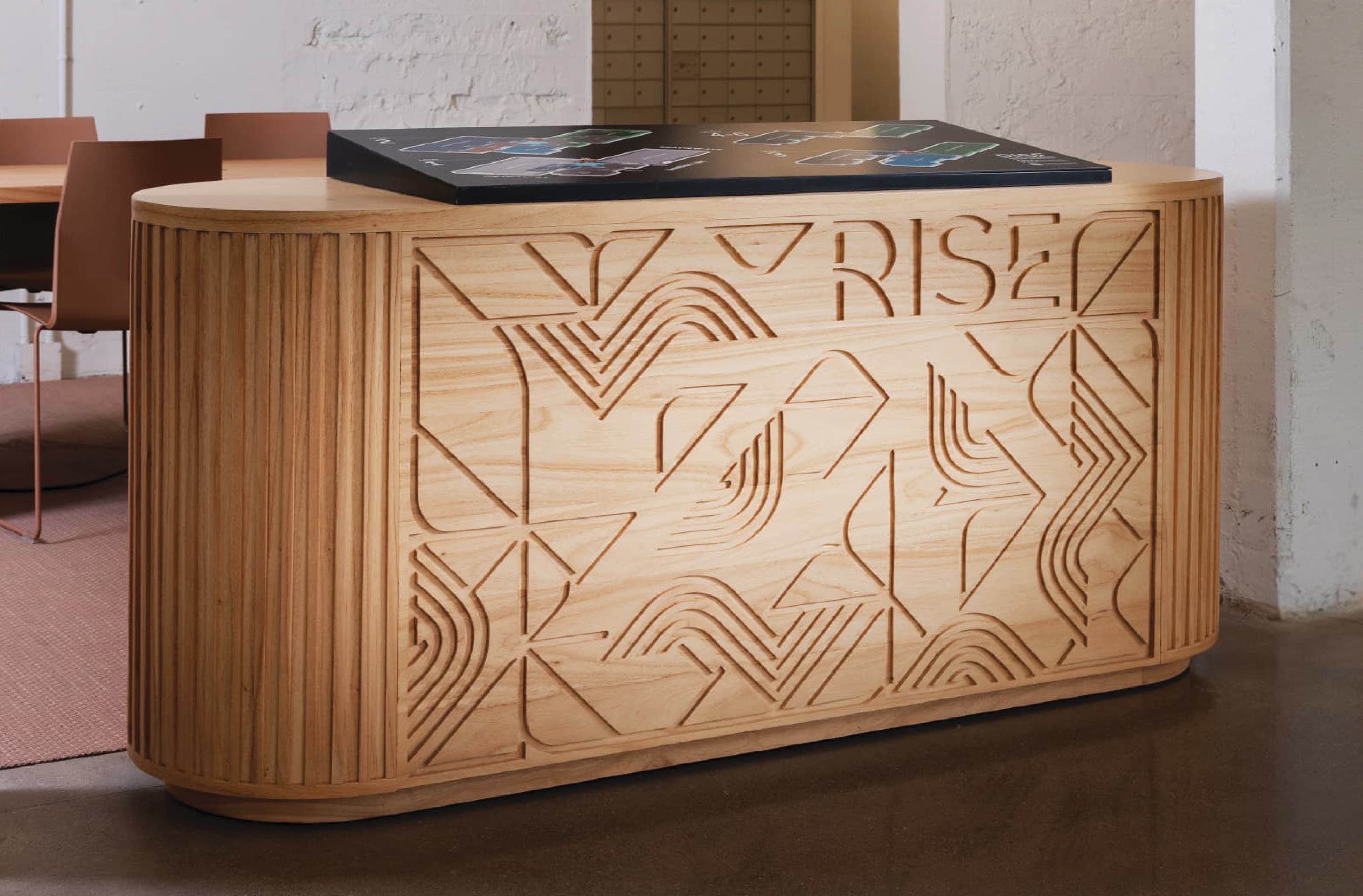 Wide view of the wooden reception desk carved with the 'RISE' logo and geometric mountain-and-wave pattern, topped by the black floor plan directory, in a lobby with terracotta chairs and a mail cubby wall in the background.