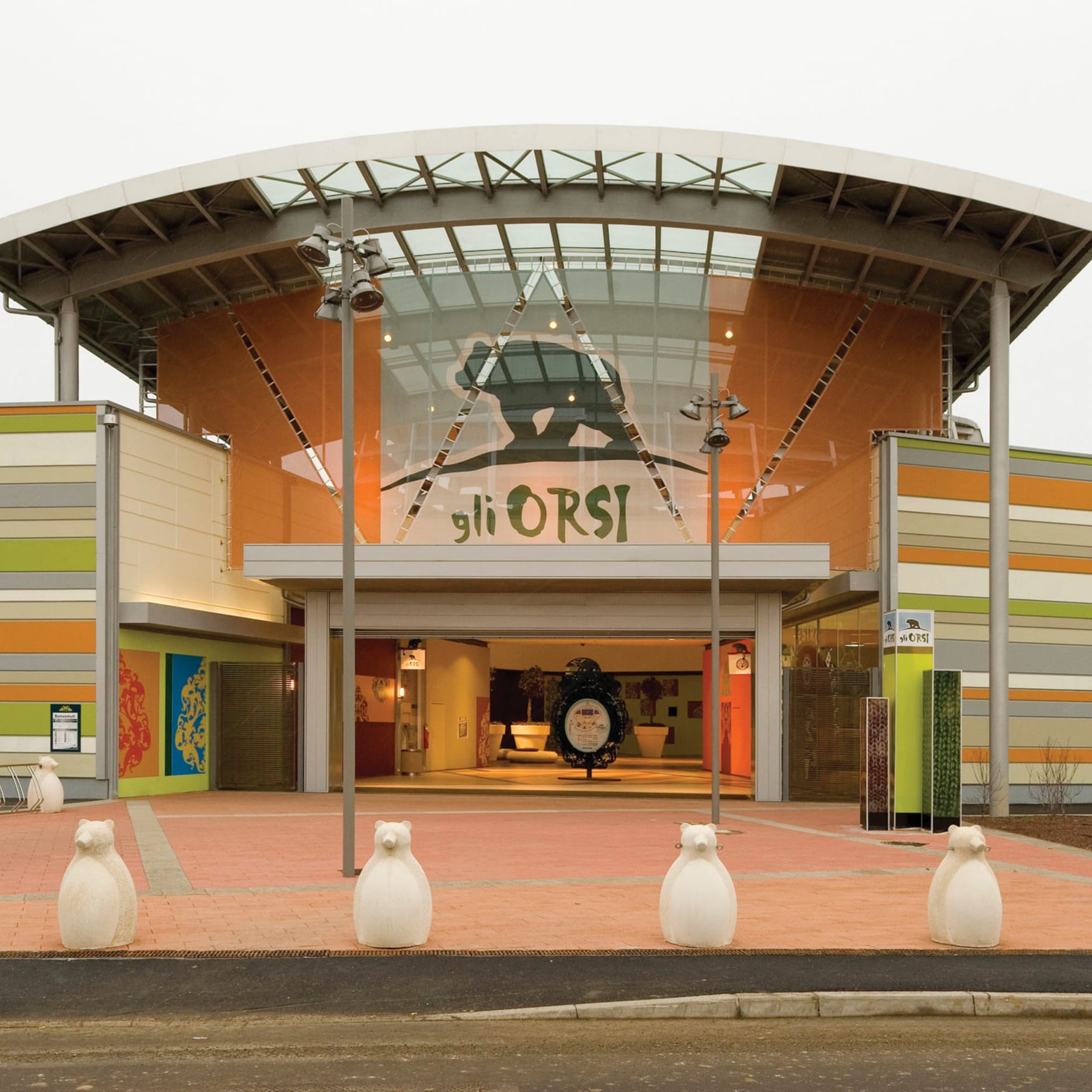 Exterior entrance of Giò Orsi featuring a modern glass atrium, colorful striped facade, sculptural bear bollards, and illuminated architectural details.