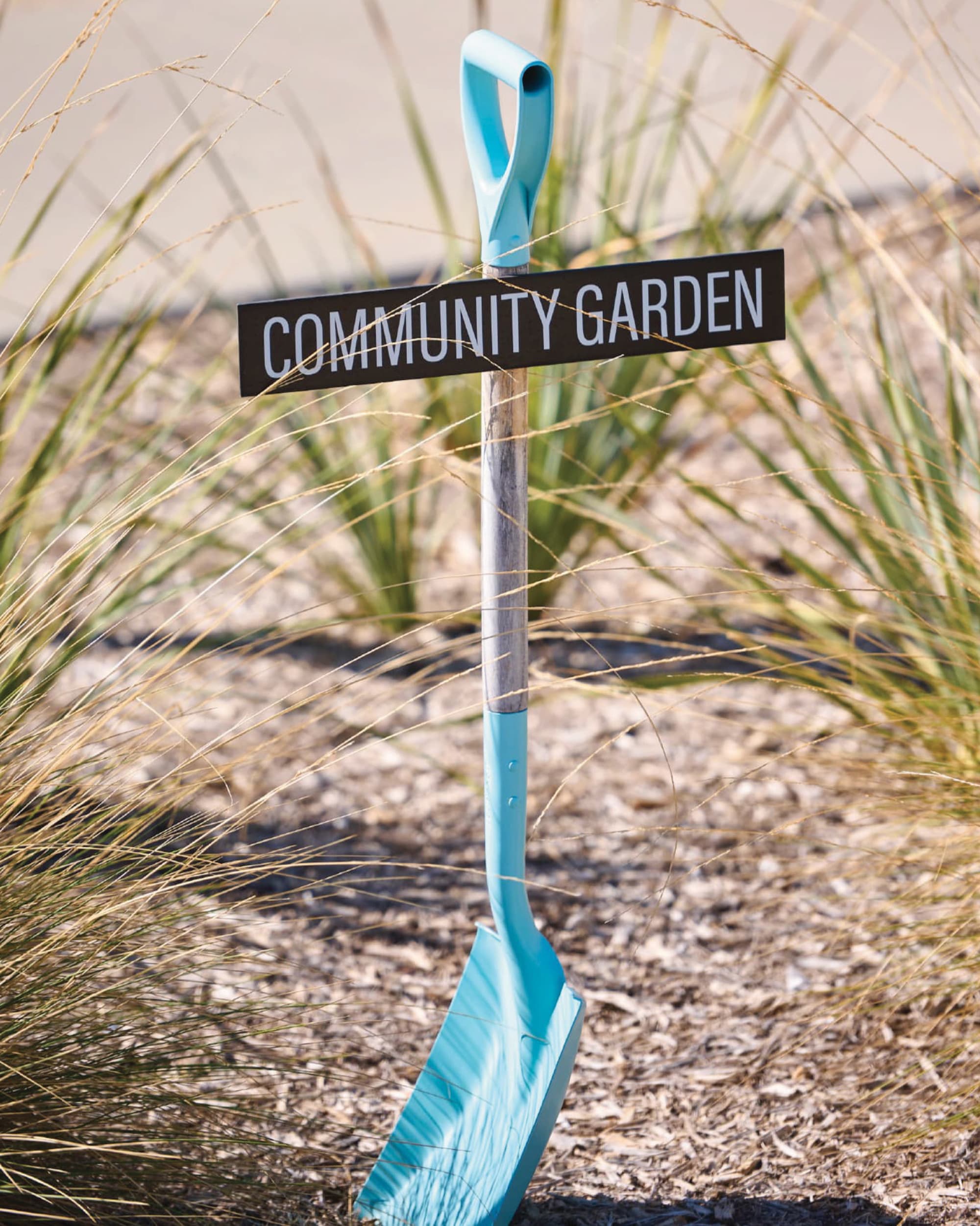 A teal shovel planted upright in the ground with a 'Community Garden' sign attached to its handle, surrounded by tall ornamental grasses.