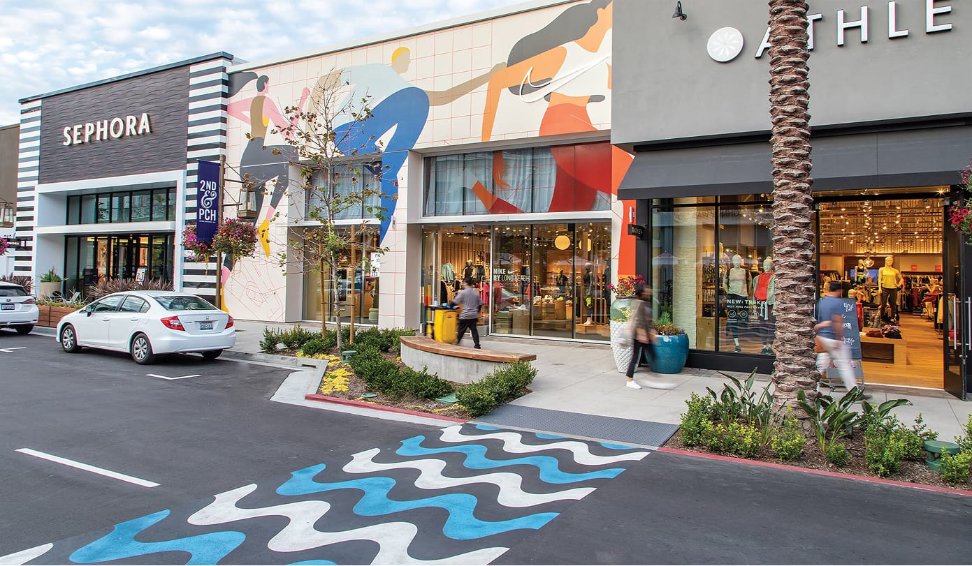 Street-level view of the retail corridor at 2nd & PCH in Long Beach, California, featuring a large-scale athletic figures mural on the Nike storefront facade, a painted wave pattern crosswalk graphic in the parking drive aisle, branded 2nd & PCH pole banner signage, and active pedestrian streetscape with Sephora and Athleta tenant storefronts — showcasing RSM Design's placemaking, experiential graphics, art programs and public art, and environmental graphic design for a coastal waterfront retail and mixed-use destination.