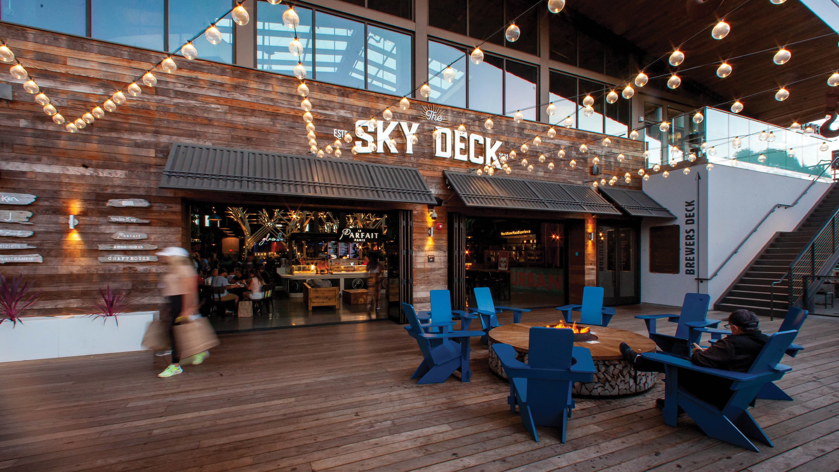 Exterior wood building facade with painted Skydeck logo, cafe lights, and a fire pit with seating