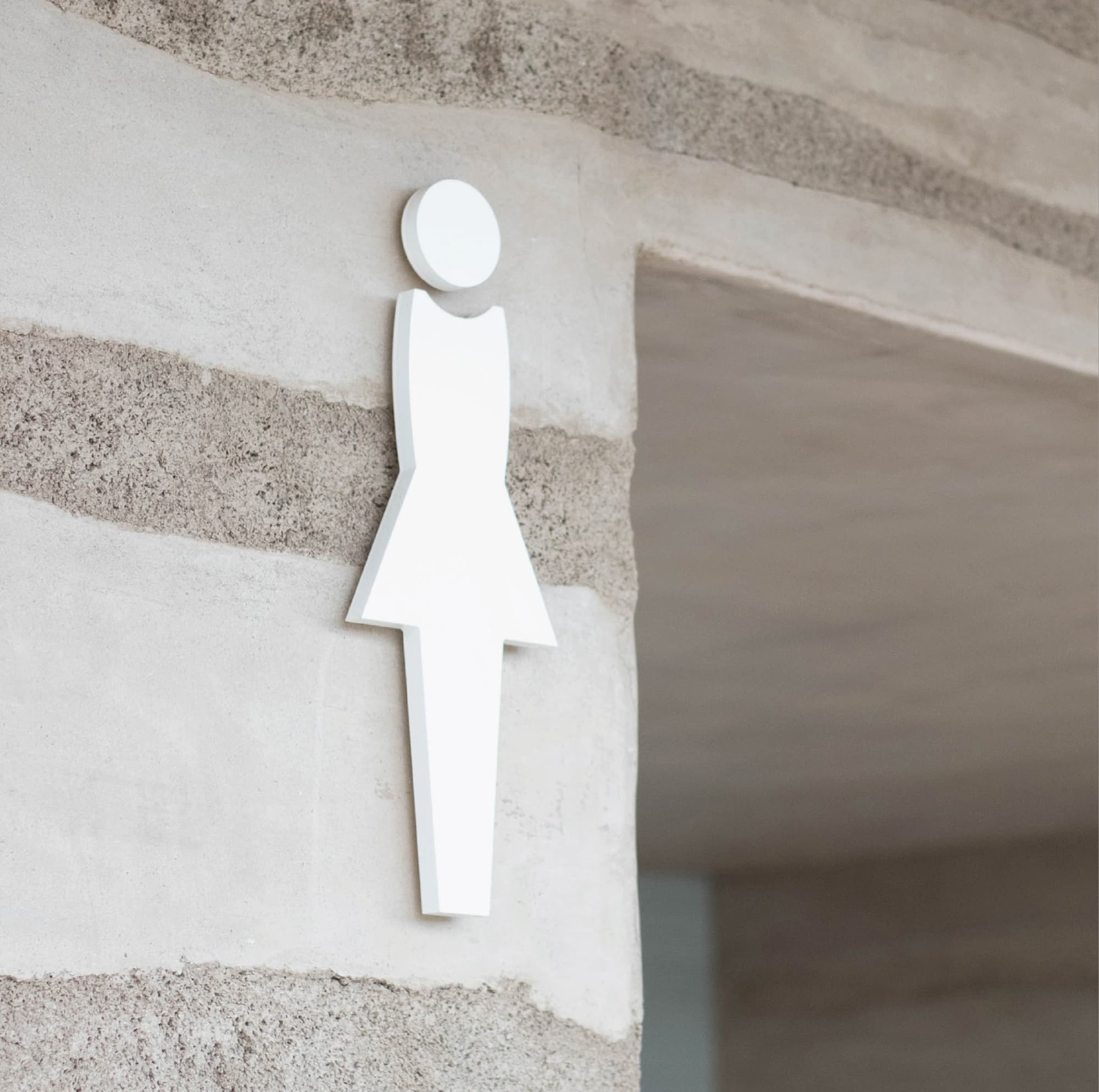 A refined dimensional white female restroom pictogram mounted on a textured rammed earth wall at the Solis Park amenity structure, its clean sculptural form complementing the natural layered concrete architecture — exemplifying RSM Design's thoughtful approach to wayfinding and signage design and specialty graphics where functional identification integrates seamlessly with architectural materials within the residential community and park markets