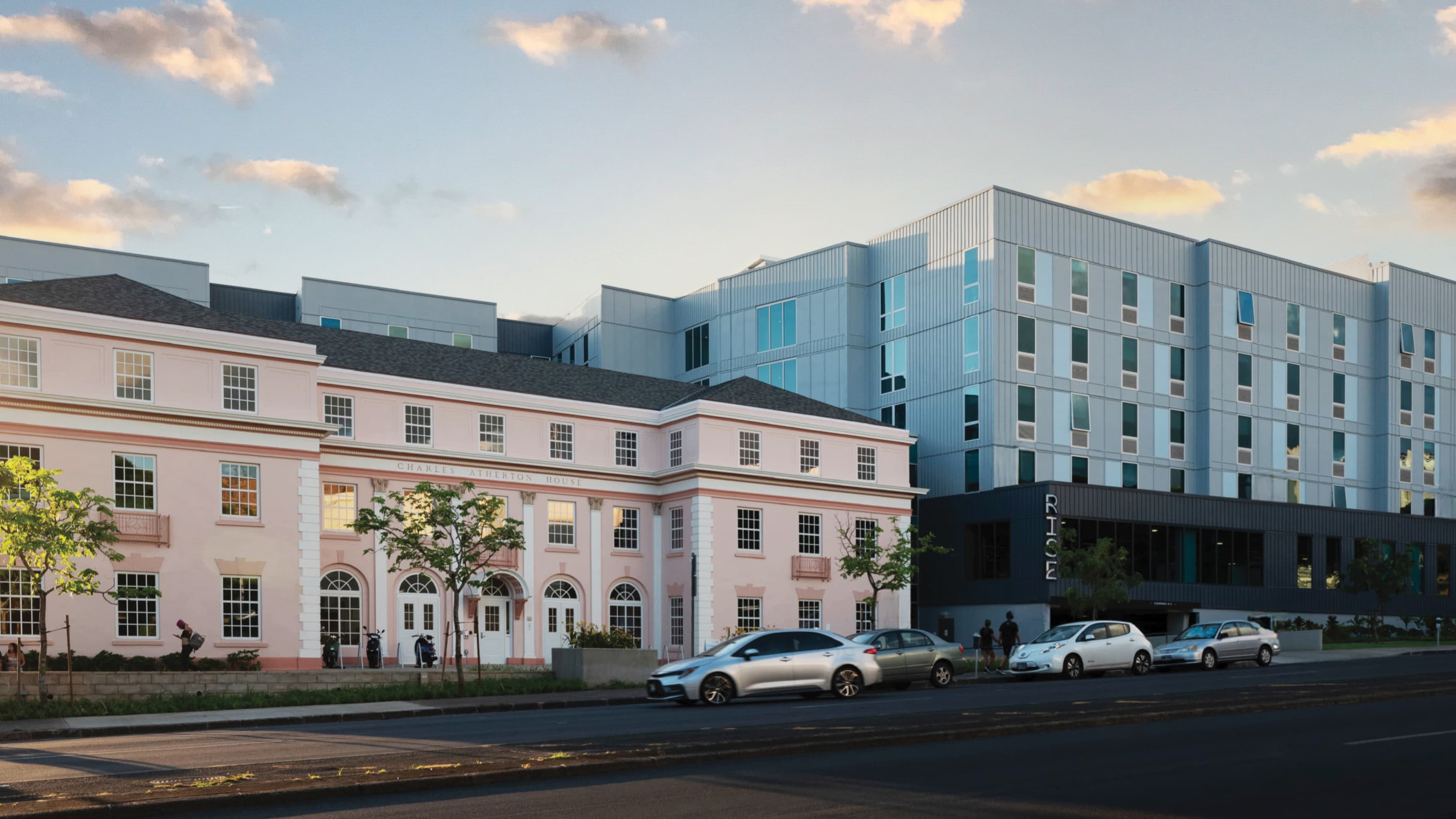 Historic pink 'Charles Atherton House' building with white columns and arched doorways, connected to a modern gray high-rise dormitory labeled 'RISE,' with cars parked along the street and pedestrians walking at dusk.