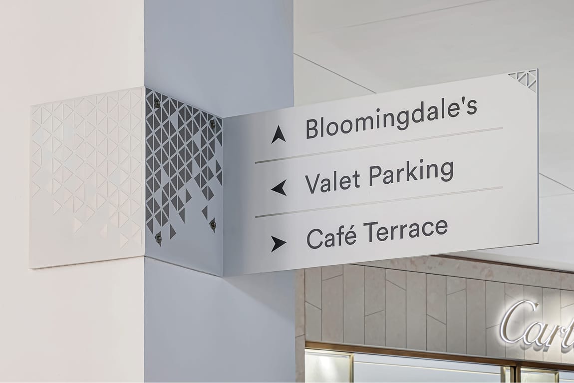 Column-mounted wayfinding sign at Fashion Valley Mall directing shoppers to Bloomingdale's, Valet Parking, and Café Terrace, with the geometric pattern bracket detail and Cartier storefront visible in the background.