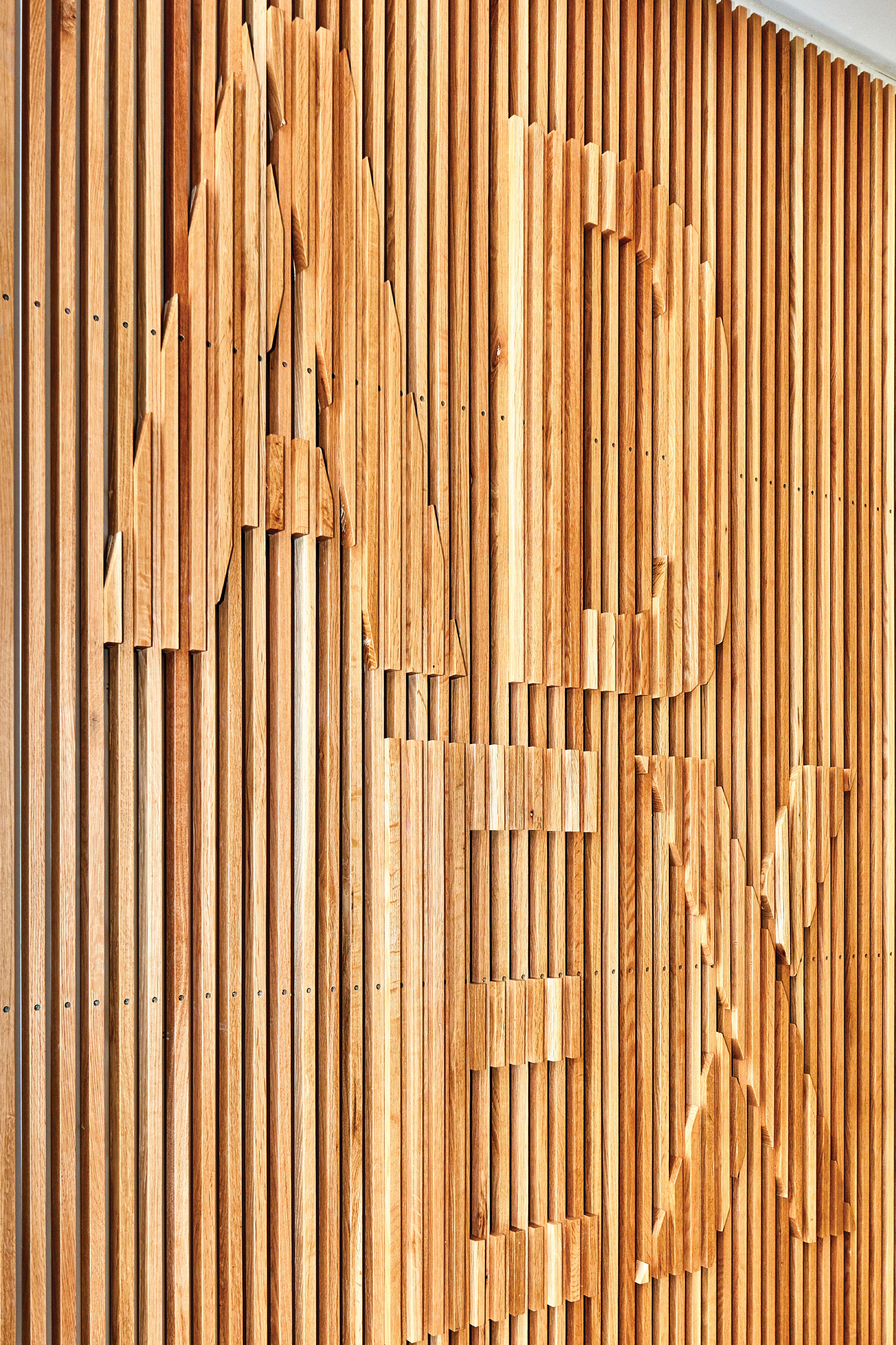 RSM Design specialty graphics and public art installation for the Architecture and Design Exchange in downtown Dallas, featuring a close-up of a large-scale dimensional wood slat wall relief with sculpted letter forms spelling ADX emerging organically from a vertical teak batten wall, demonstrating how environmental graphic design, art programs, experiential graphics, and placemaking transform interior architectural surfaces into memorable, craft-driven branded environments for cultural, workplace, and civic markets.