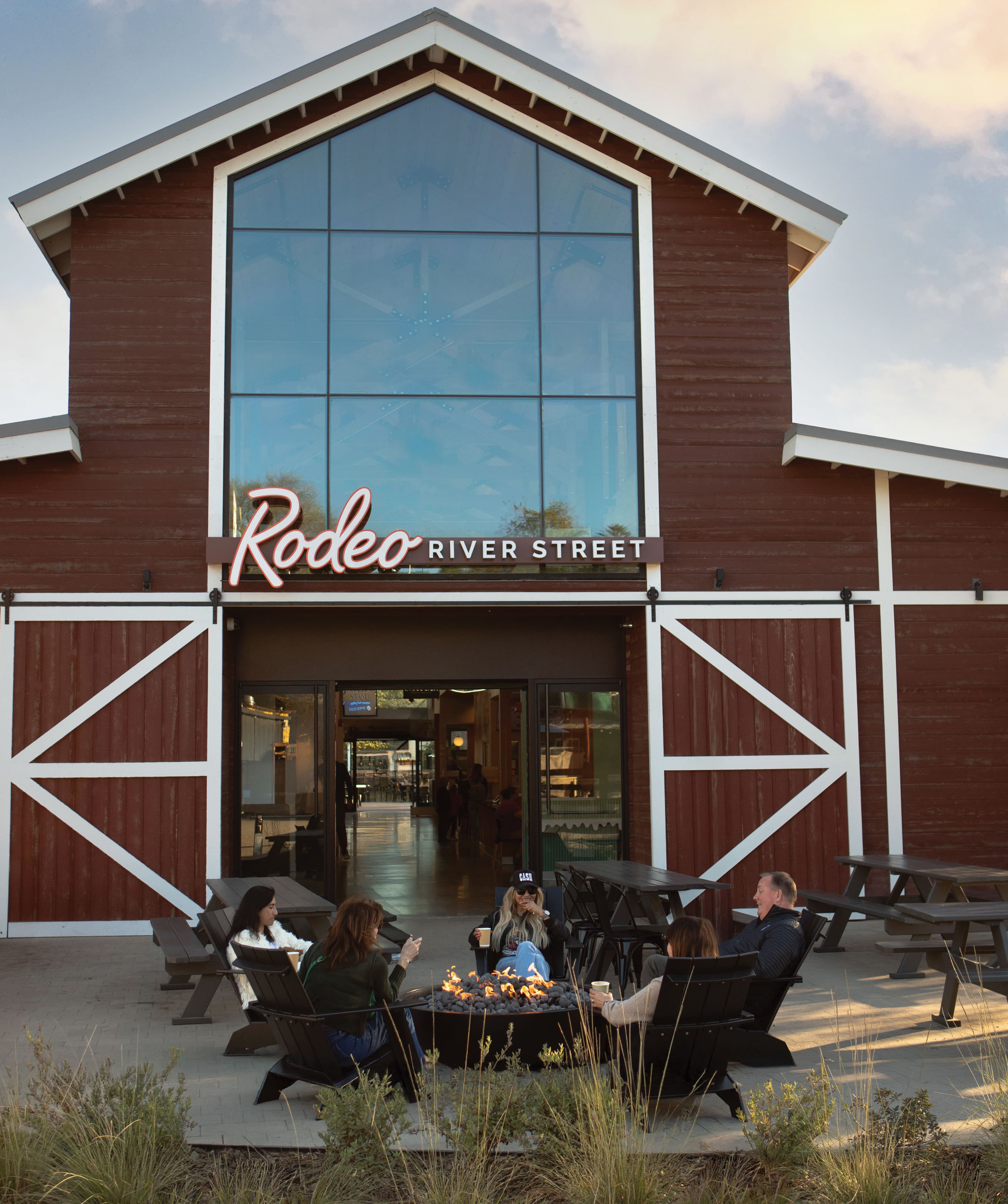 RSM Design entry signage and placemaking design for Rodeo River Street, featuring an illuminated script and serif blade sign mounted above barn-style sliding doors on a red wood-clad building facade, with people gathered around a fire pit in the foreground, illustrating how environmental graphic design and branded environments create human-centered gathering places for food and beverage, mixed-use, and experiential retail markets.