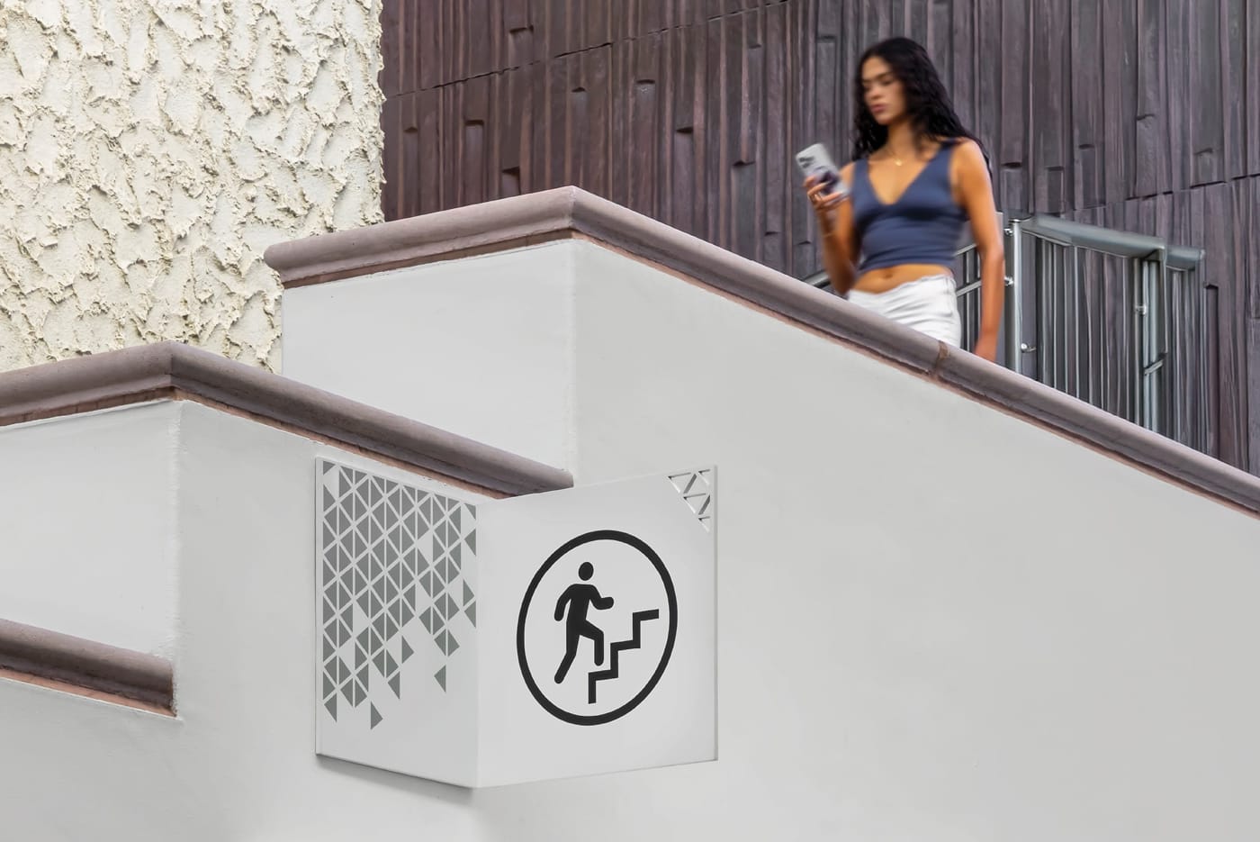 Close-up of a Fashion Valley Mall staircase wayfinding sign featuring a geometric pattern panel and circular stair icon, with a shopper descending the stairs in the background.