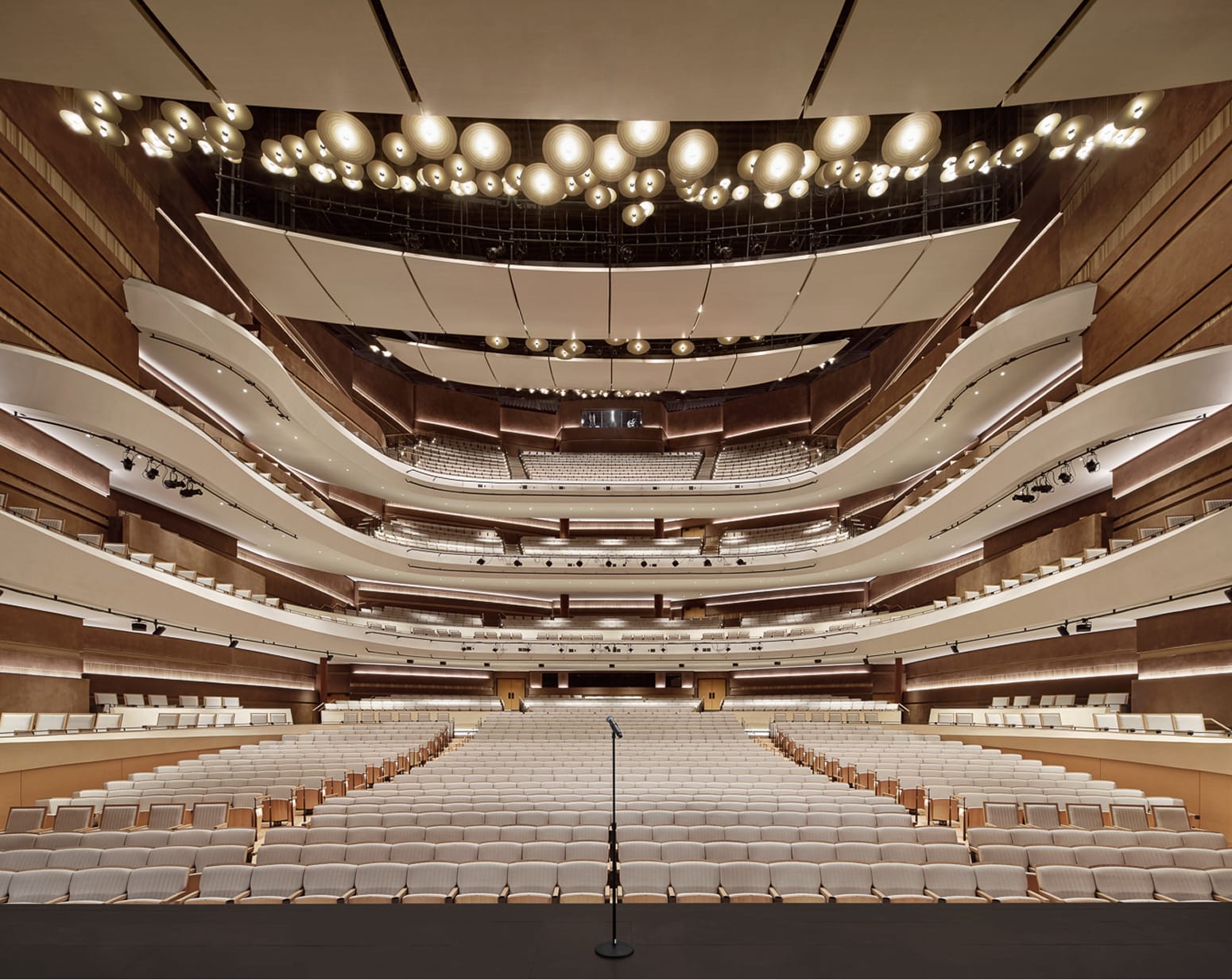 View from the stage of the grand multi-tiered concert hall auditorium at Buddy Holly Hall of Performing Arts and Sciences, featuring sweeping curved balconies, warm wood paneling, globe pendant lighting, and over 2,200 seats, Lubbock Texas