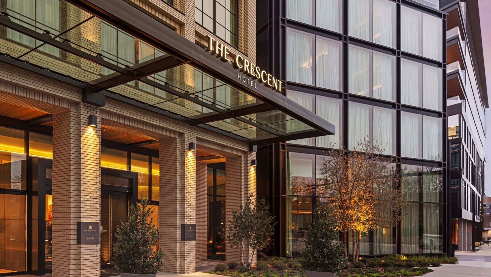 The Crescent Hotel exterior entrance at dusk featuring illuminated glass canopy, warm-lit brick facade, manicured landscaping with uplighted trees, and floor-to-ceiling windows of an upscale boutique hotel