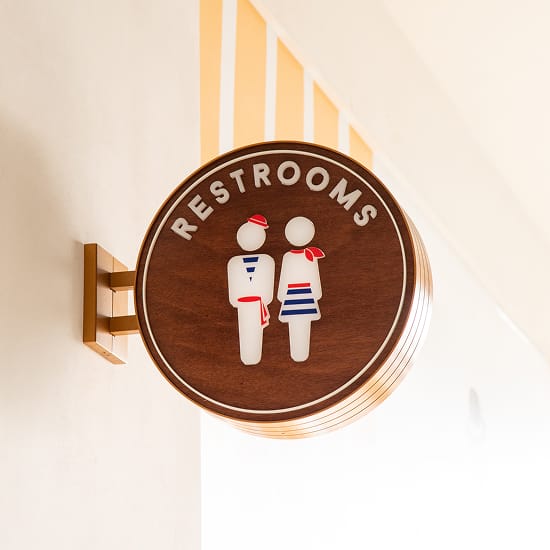 Branded circular projecting restroom identification sign at 2nd & PCH in Long Beach, California, featuring a walnut wood face with a brass frame, dimensional 'Restrooms' lettering, and playful nautical-themed pictogram figures dressed as sailors — an example of RSM Design's wayfinding signage design and specialty graphic design that brings moments of delight to a coastal waterfront retail and mixed-use destination.