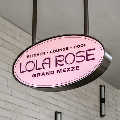 Illuminated oval blade sign for Lola Rose Grand Mezze — Kitchen, Lounge, Pool — in deep plum typography on a pink background with a dark metal frame, suspended from the ceiling against a white brick wall.