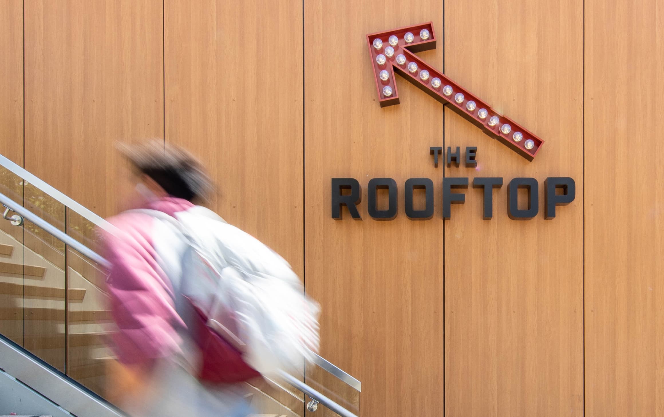 Red directional open channel arrow with exposed light bulbs mounted wall by staircase above black "THE ROOFTOP" dimensional letters signage for UC San Diego Sixth College Dining Hall by RSM Design in San Diego, California.