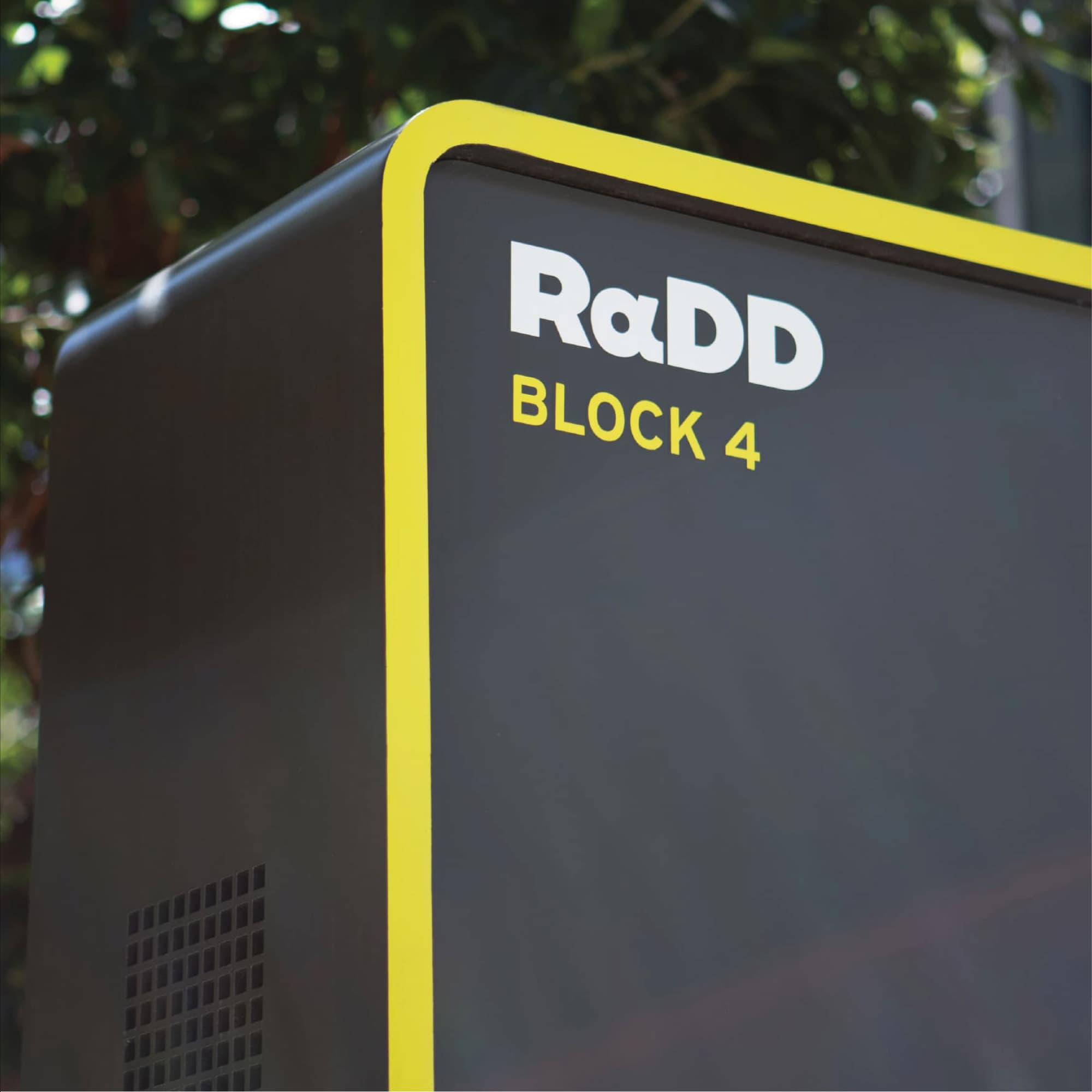 Close-up of a dark gray monument sign with a yellow edge border, reading 'RαDD Block 4,' set against blurred trees in the background.