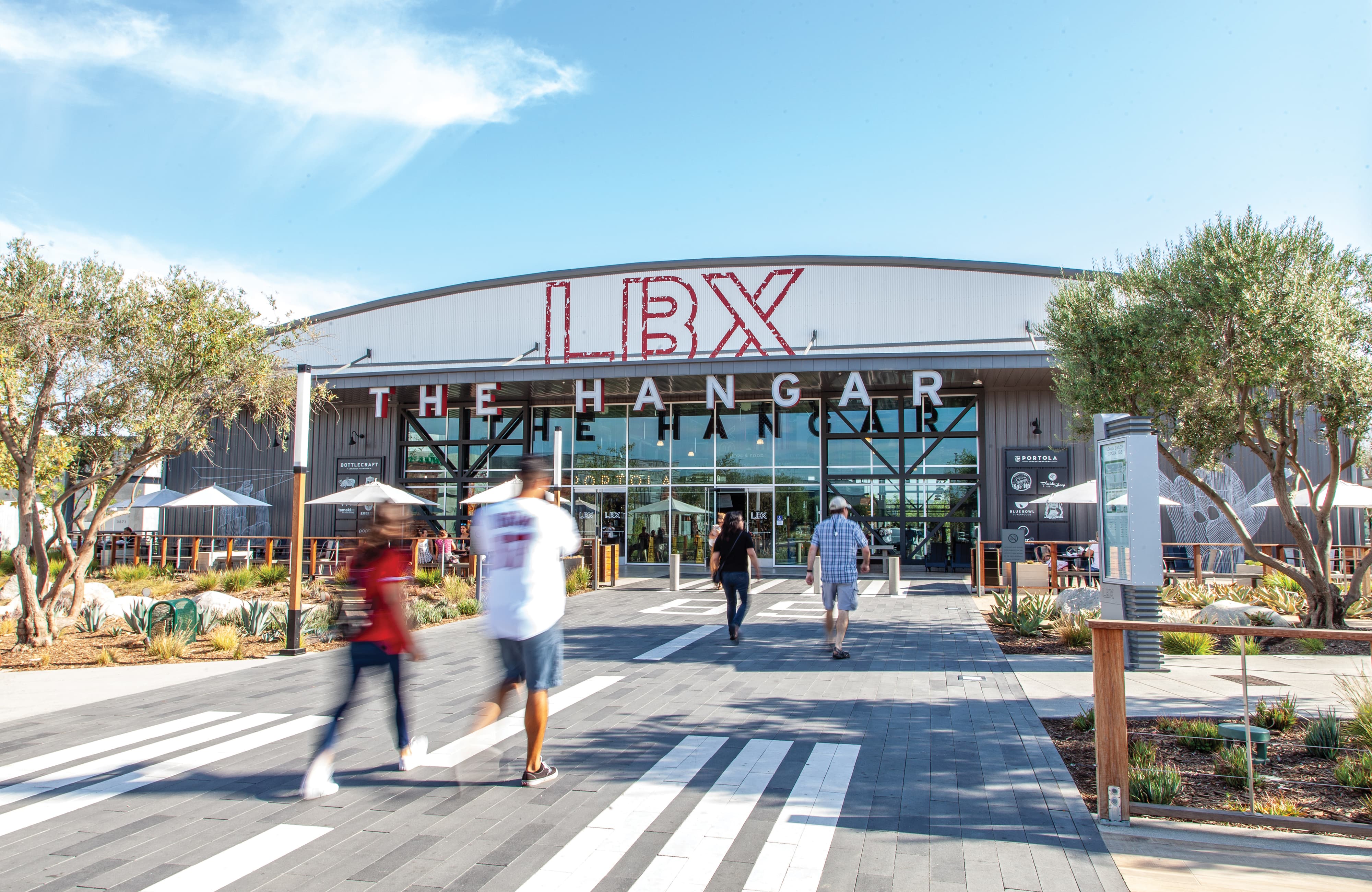 Exterior of 'LBX, The Hangar' retail building with a curved metal roof, glass storefront entrance, and storefront signage including Bottlecraft and Portola, as pedestrians cross a paved plaza dotted with olive trees and desert landscaping.
