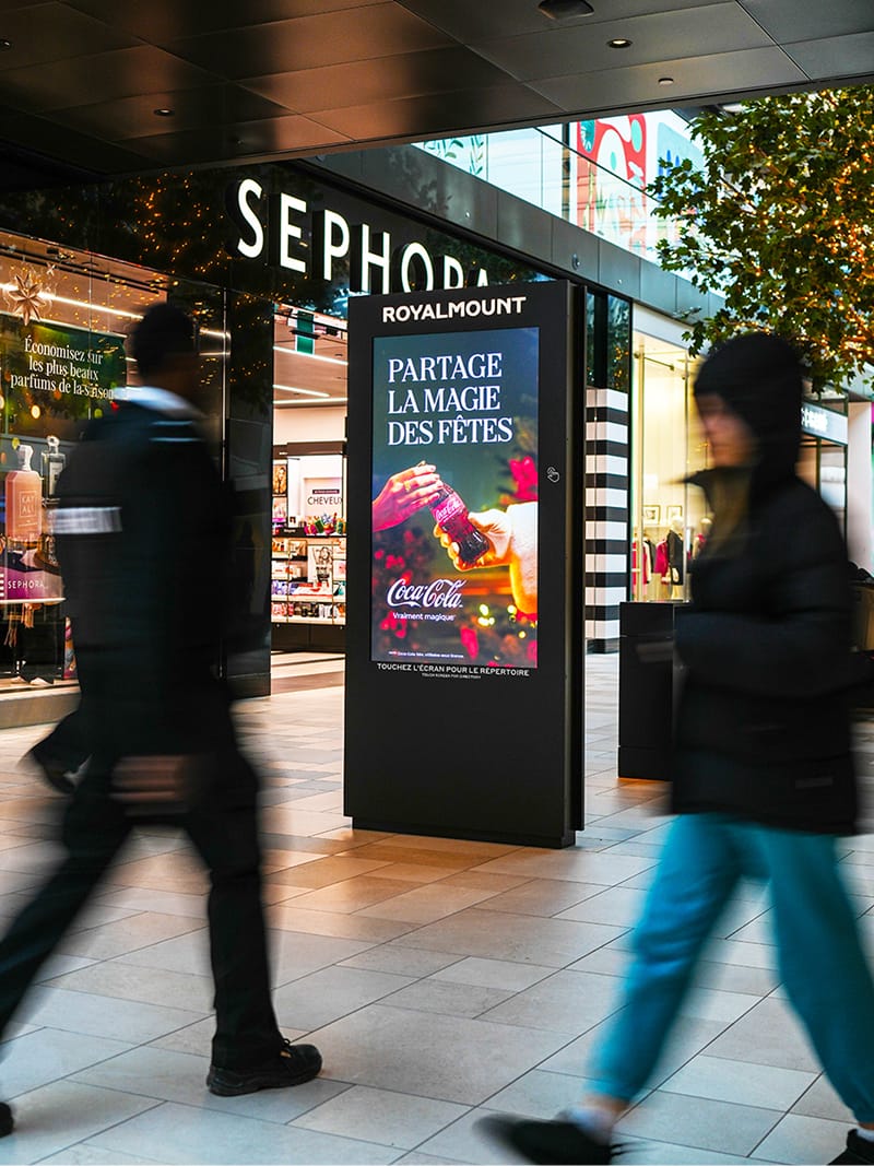Royalmount digital advertising column with holiday-themed Coca-Cola display and touch-to-browse directory prompt, shoppers passing by Sephora storefront — digital signage design by RSM Design