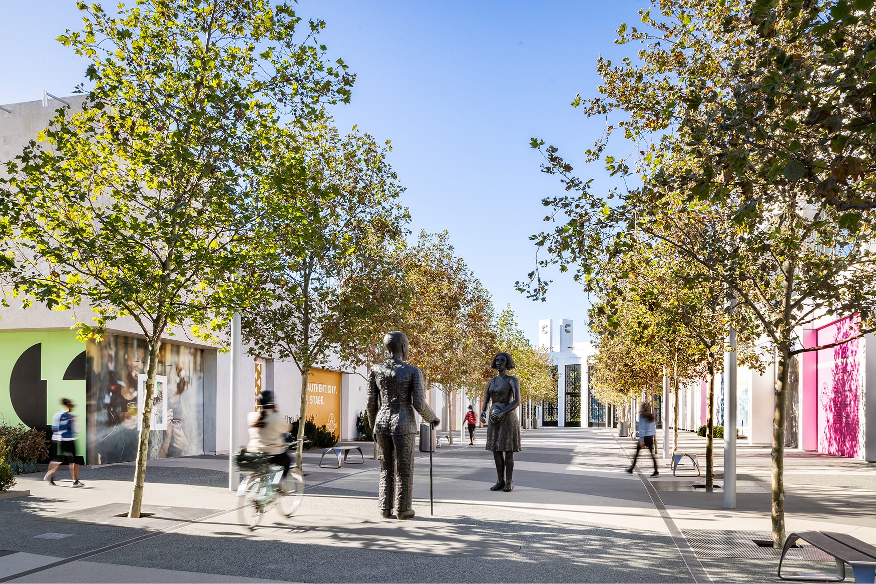 Hollywood Park pedestrian promenade with bronze figurative sculptures, tree-lined walkway, colorful retail storefronts, and shoppers on sunny day in Inglewood, California
