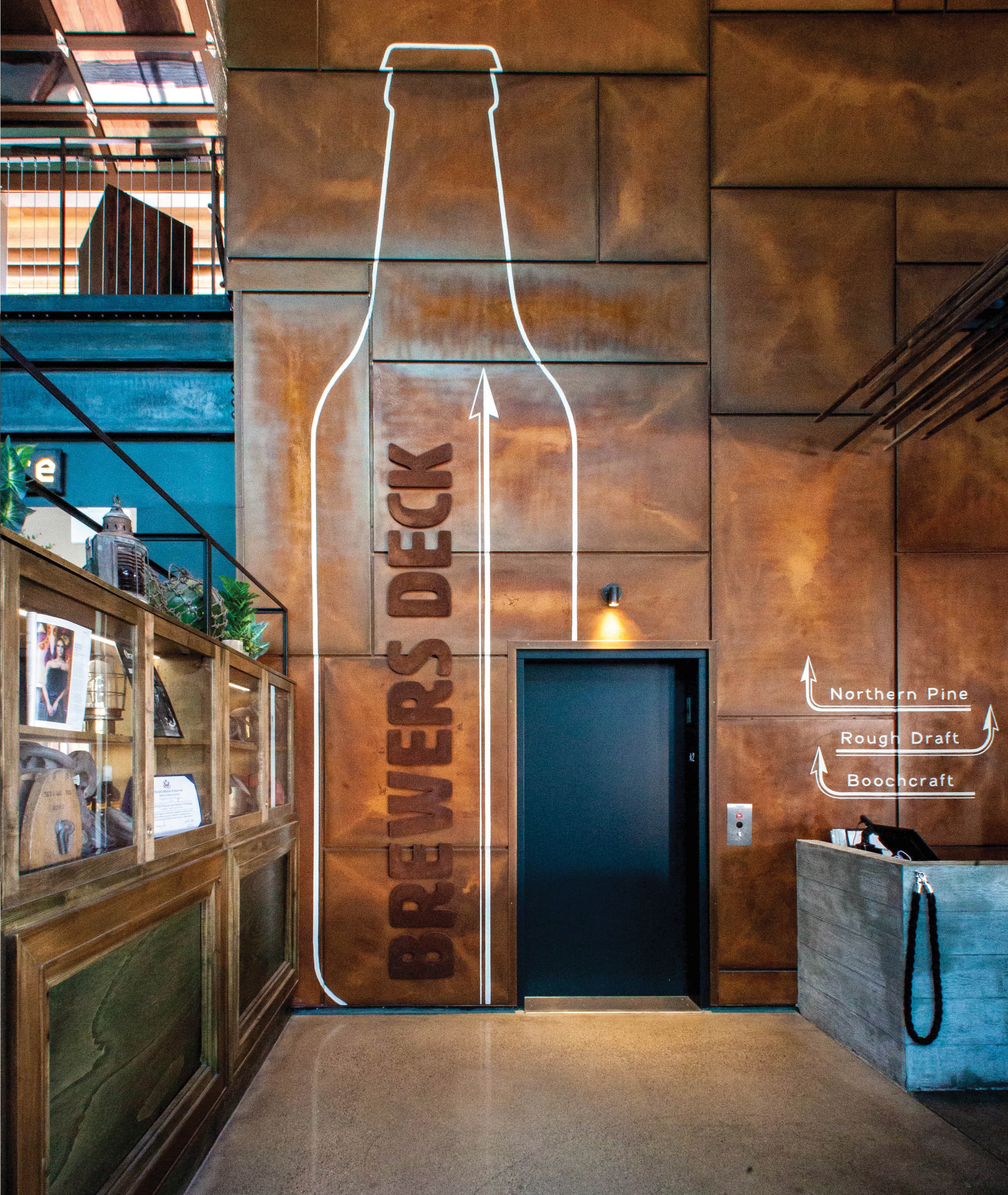 Interior entry wall of Crafthouse Sky Deck featuring a floor-to-ceiling neon bottle outline with a dimensional "Brewers Deck" typographic directional arrow on weathered copper wall panels, flanked by hand-lettered wayfinding arrows directing guests to Northern Pine, Rough Draft, and Boochcraft — demonstrating RSM Design's integrated approach to experiential graphics and branded environments, specialty graphics, and wayfinding and signage design within the food and beverage and mixed-use markets