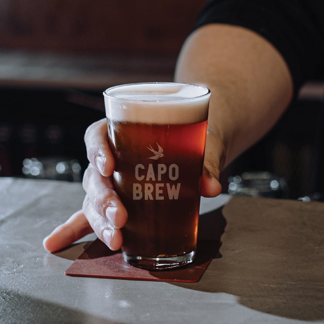 Hand holding an etched Capo Brew pint glass filled with amber beer at a bar, brand identity design by RSM Design