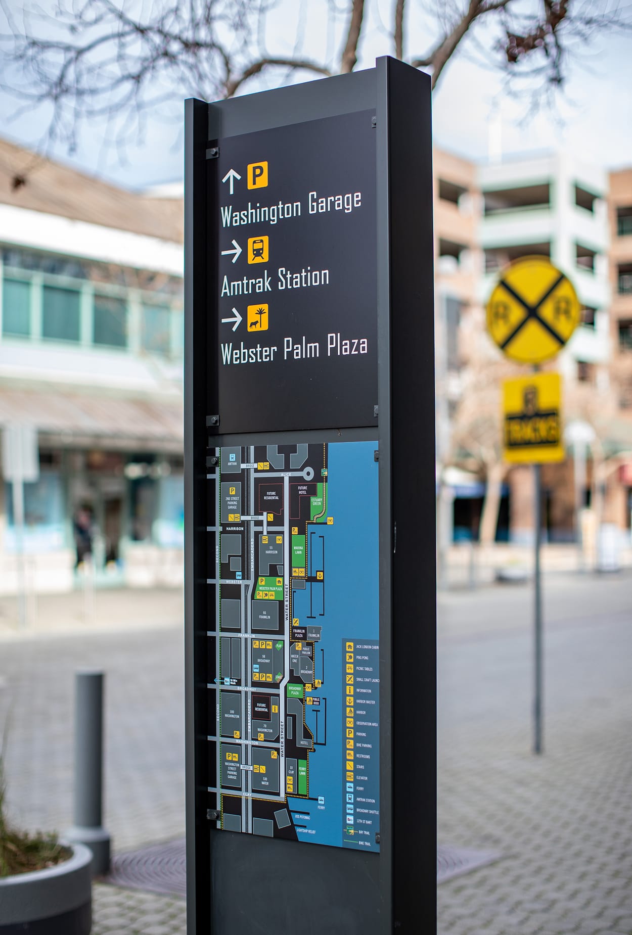 Jack London Square wayfinding kiosk sign with directional arrows to Washington Garage parking, Amtrak Station, and Webster Palm Plaza, with neighborhood map panel and railroad crossing sign in background, Oakland, California