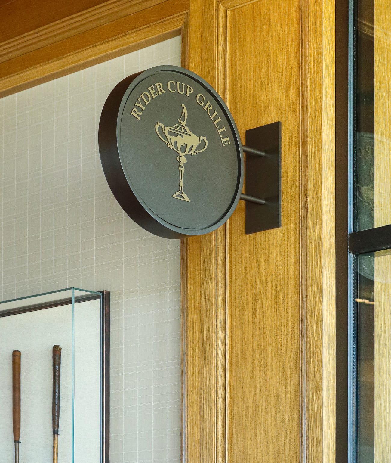 Ryder Cup Grille circular projecting sign with gold trophy illustration mounted on warm oak wood paneling at Omni PGA Frisco, with vintage golf clubs displayed in glass case nearby