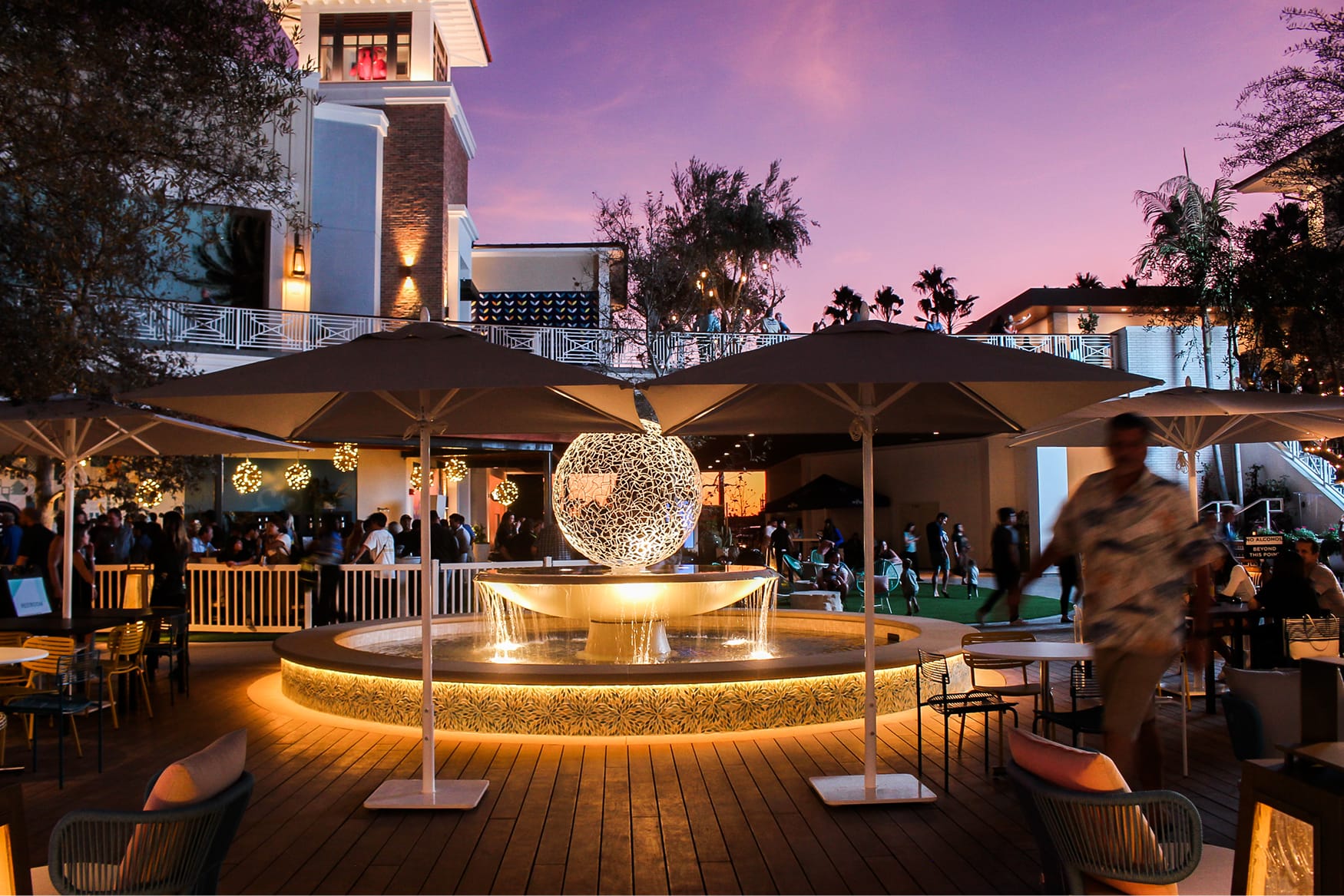 Dusk view of the activated outdoor plaza at 2nd & PCH in Long Beach, California, centered on an illuminated sculptural orb fountain surrounded by an animated crowd, outdoor dining, and a floral mosaic-tiled fountain base beneath a purple sunset sky — showcasing RSM Design's placemaking, art programs and public art, and experiential graphic design that connects people to place within a coastal waterfront retail and mixed-use destination.