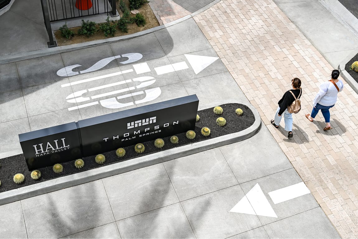 Aerial view of the Thompson Palm Springs and Hall Napa Valley monument sign surrounded by golden barrel cacti, with two guests walking along an adjacent brick pathway and bold graphic wayfinding arrows painted on the concrete drive.