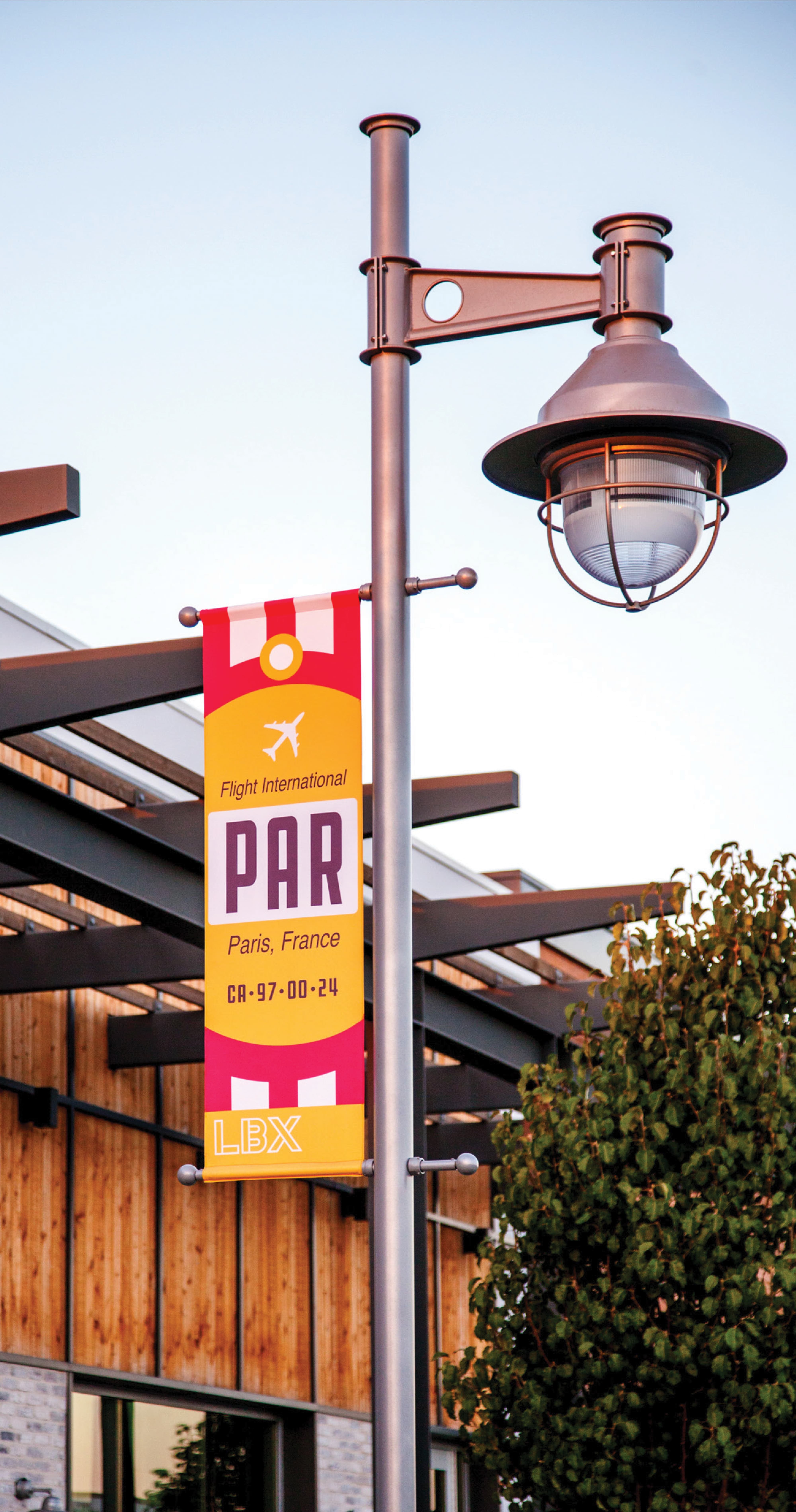 Vintage airline-ticket style banner reading 'Flight International, PAR, Paris, France, LBX' hanging from an ornate lamppost with a caged glass light fixture, in front of a wood-slat building and tree at dusk.