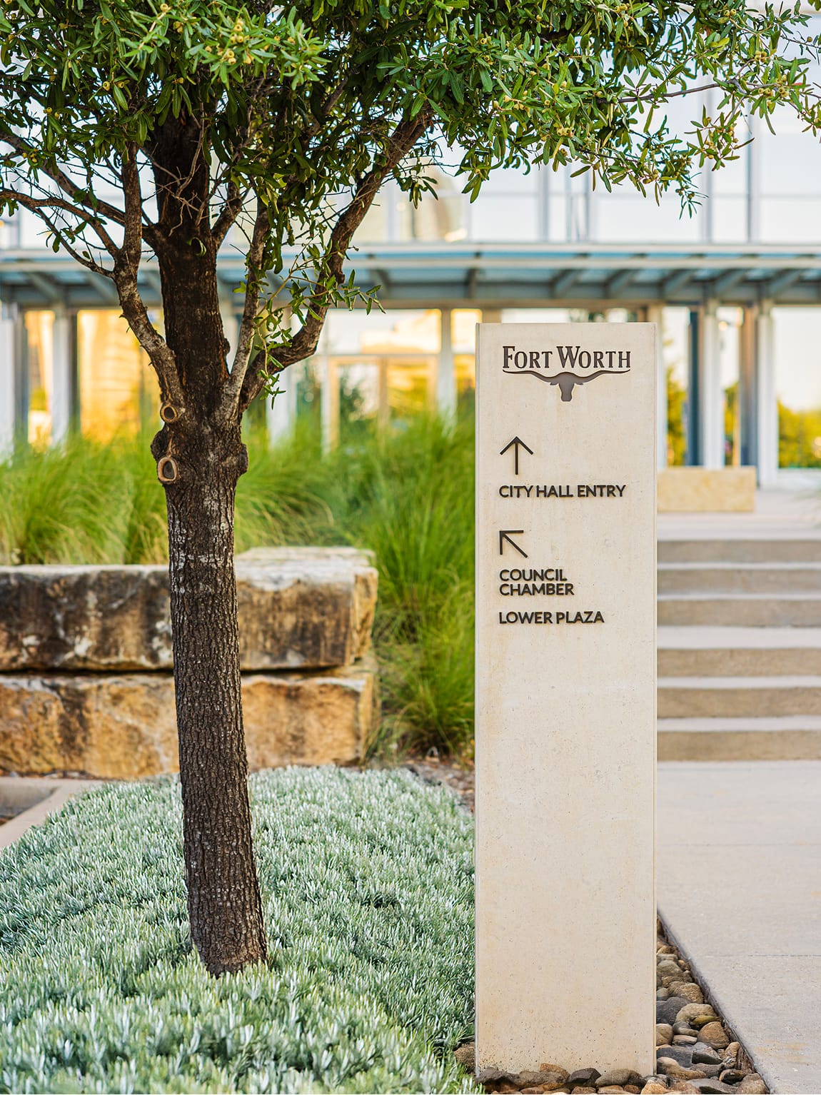Fort Worth City Hall exterior wayfinding bollard in limestone finish with longhorn logo directing visitors to City Hall Entry, Council Chamber, and Lower Plaza amid native landscaping and stone retaining walls