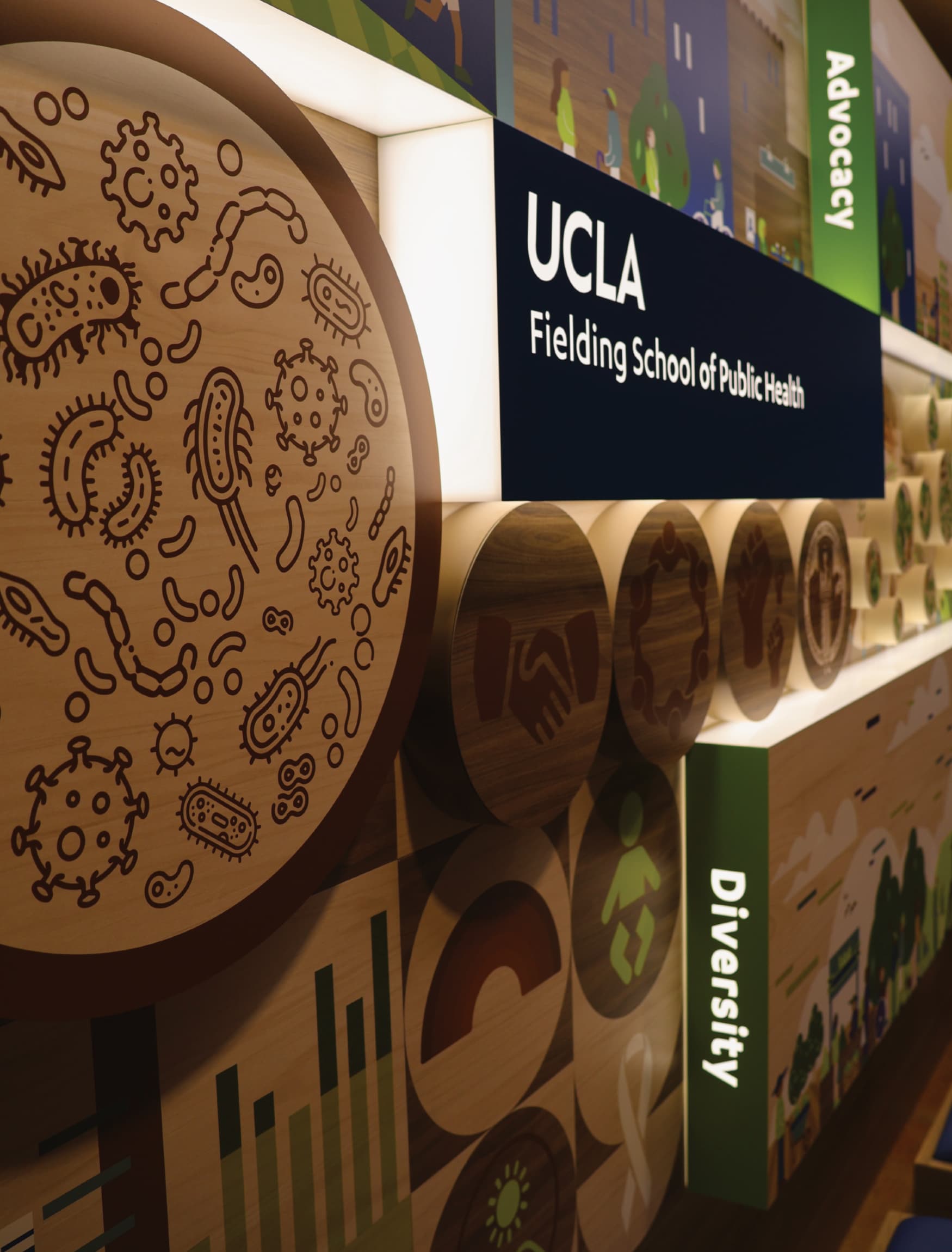 The wall in the image is part of the permanent exhibition designed by RSM Design for the UCLA Fielding School of Public Health's 60th anniversary. It features a vibrant and informative display with a variety of graphic elements and icons. Key public health themes such as Inclusion, Equity, Evidence, Prevention, Community, and Impact are highlighted on the wall, each with its own distinct section. The design includes detailed illustrations, representing different aspects of public health and the school's contributions to the field. The wall is well-lit and incorporates wooden textures with colorful visual elements, making the information engaging and accessible. There are also seating benches integrated into the design, inviting viewers to spend time interacting with the exhibit.