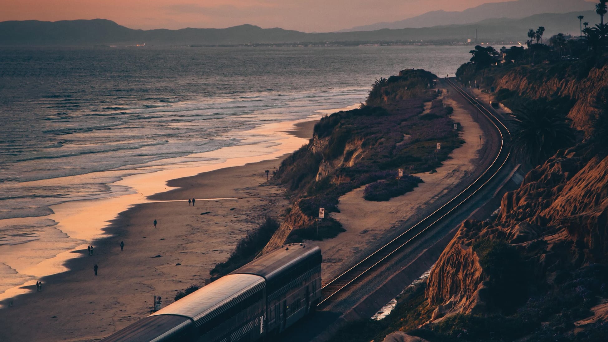 Elevated view of a passenger train traveling along coastal railroad tracks atop bluffs above a sandy beach, with people walking along the shore and a distant mountain range at dusk.