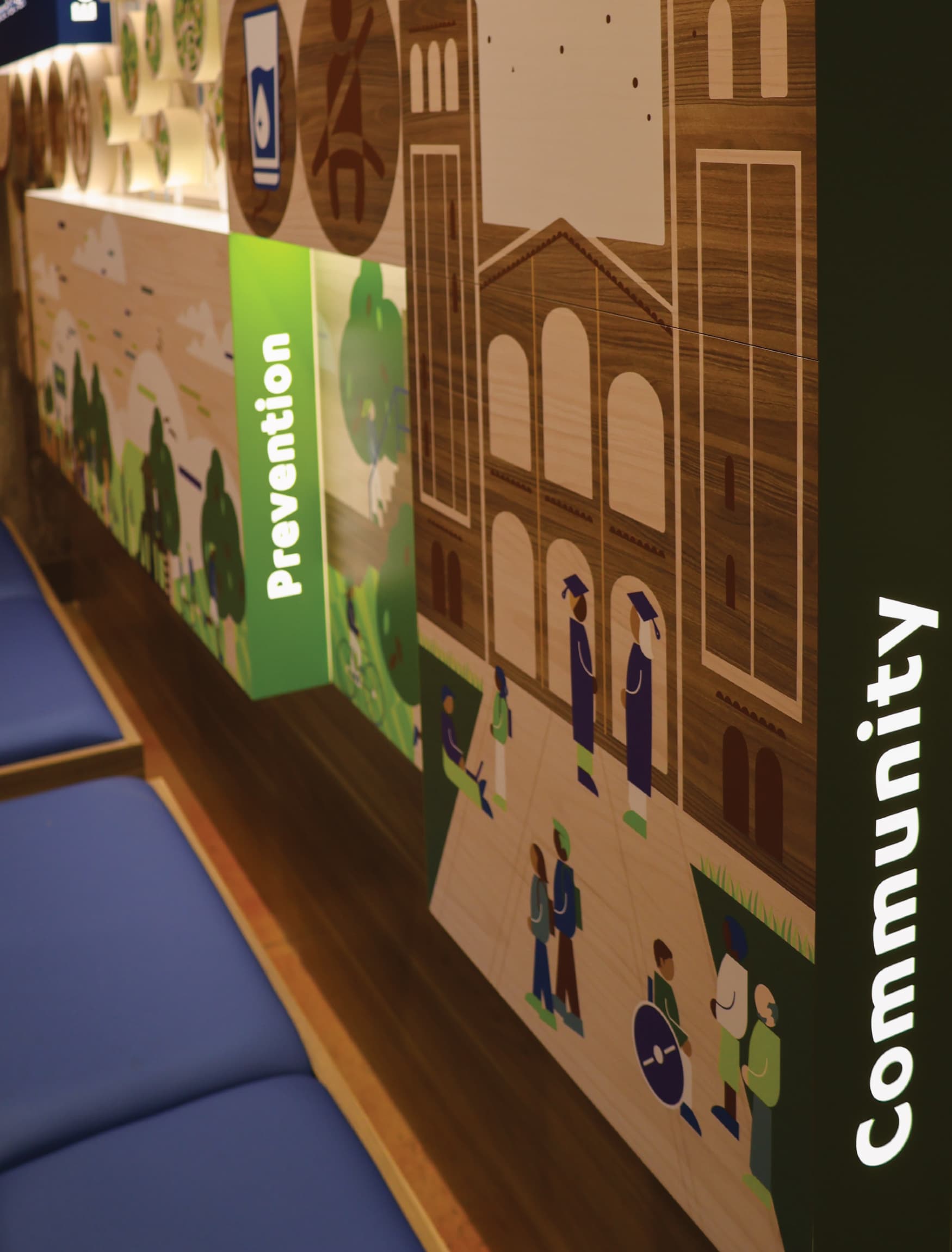 The wall in the image is part of the permanent exhibition designed by RSM Design for the UCLA Fielding School of Public Health's 60th anniversary. It features a vibrant and informative display with a variety of graphic elements and icons. Key public health themes such as Inclusion, Equity, Evidence, Prevention, Community, and Impact are highlighted on the wall, each with its own distinct section. The design includes detailed illustrations, representing different aspects of public health and the school's contributions to the field. The wall is well-lit and incorporates wooden textures with colorful visual elements, making the information engaging and accessible. There are also seating benches integrated into the design, inviting viewers to spend time interacting with the exhibit.