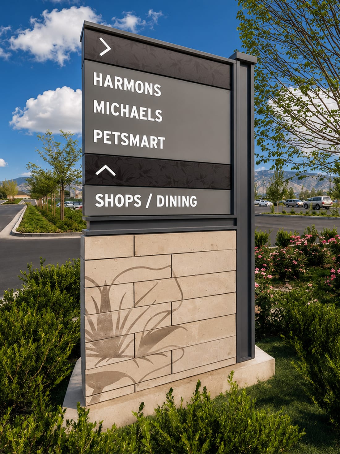 Directional wayfinding sign listing Harmons, Michaels, and PetSmart with directional arrows to Shops/Dining, set on a stone base etched with a sego lily motif at Mountain View Village in Riverton, Utah — wayfinding and environmental graphic design by RSM Design.