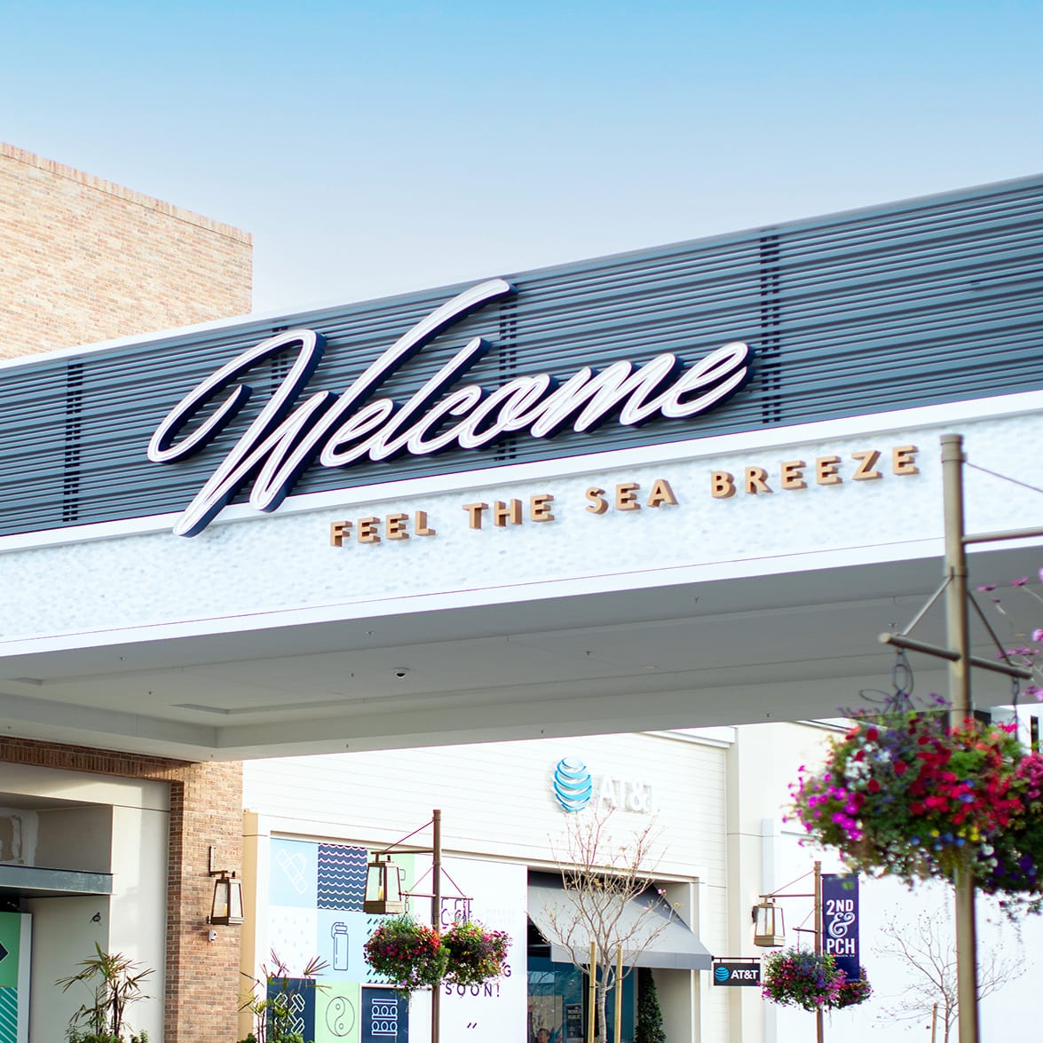 Exterior entrance gateway at 2nd & PCH in Long Beach, California, featuring large-scale dimensional script 'Welcome' signage and 'Feel the Sea Breeze' tagline lettering mounted on a pedestrian breezeway, with branded 2nd & PCH banner signage and tenant storefronts visible below — showcasing RSM Design's placemaking, experiential graphics, and branded environments for a coastal waterfront retail and mixed-use destination.