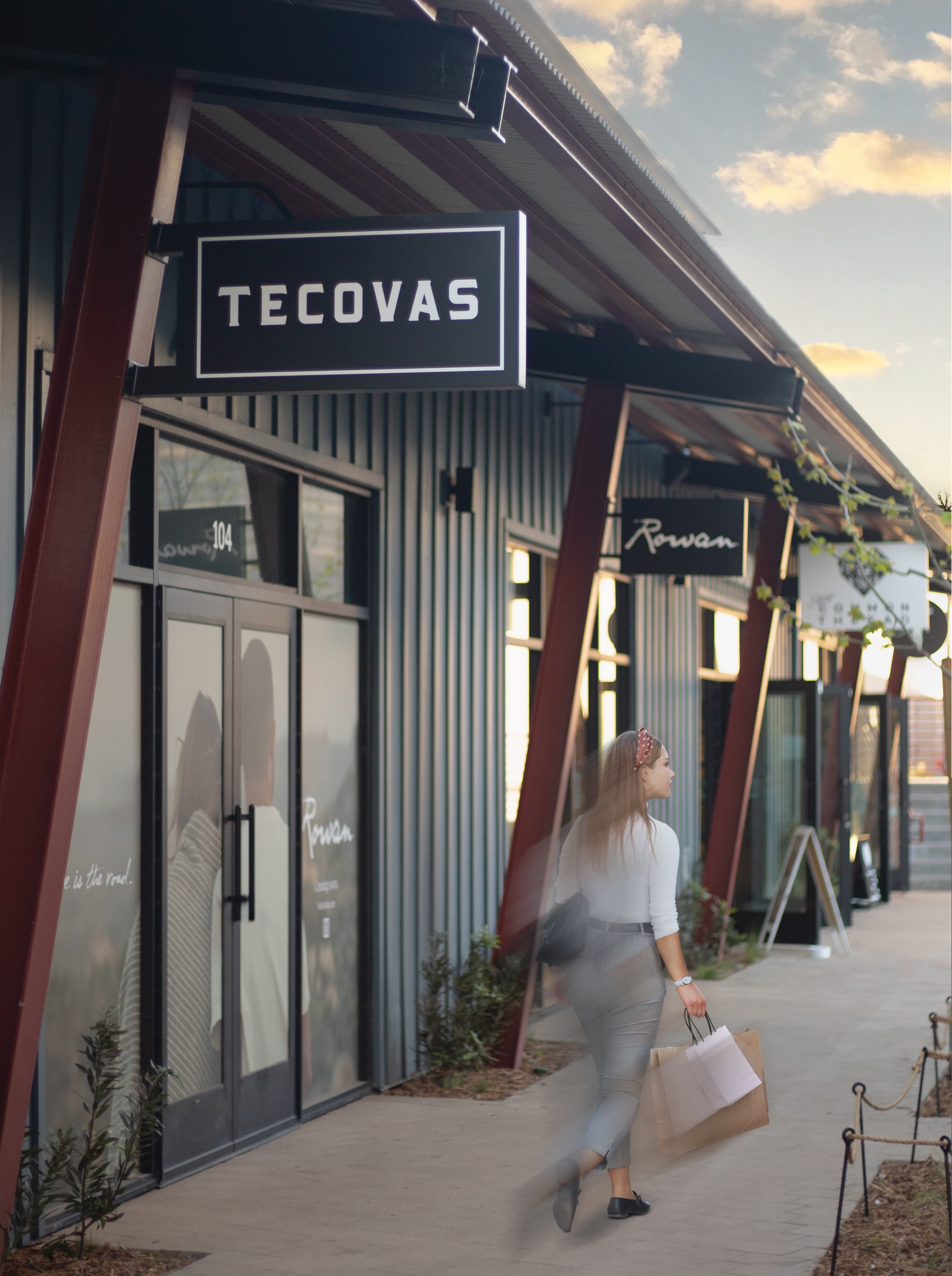 RSM Design wayfinding and signage design for River Street Marketplace, featuring illuminated tenant blade signs for retail storefronts including Tecovas and Rowan, integrated into a mixed-use streetscape environment where a pedestrian walks along a well-branded, human-centered outdoor retail corridor.