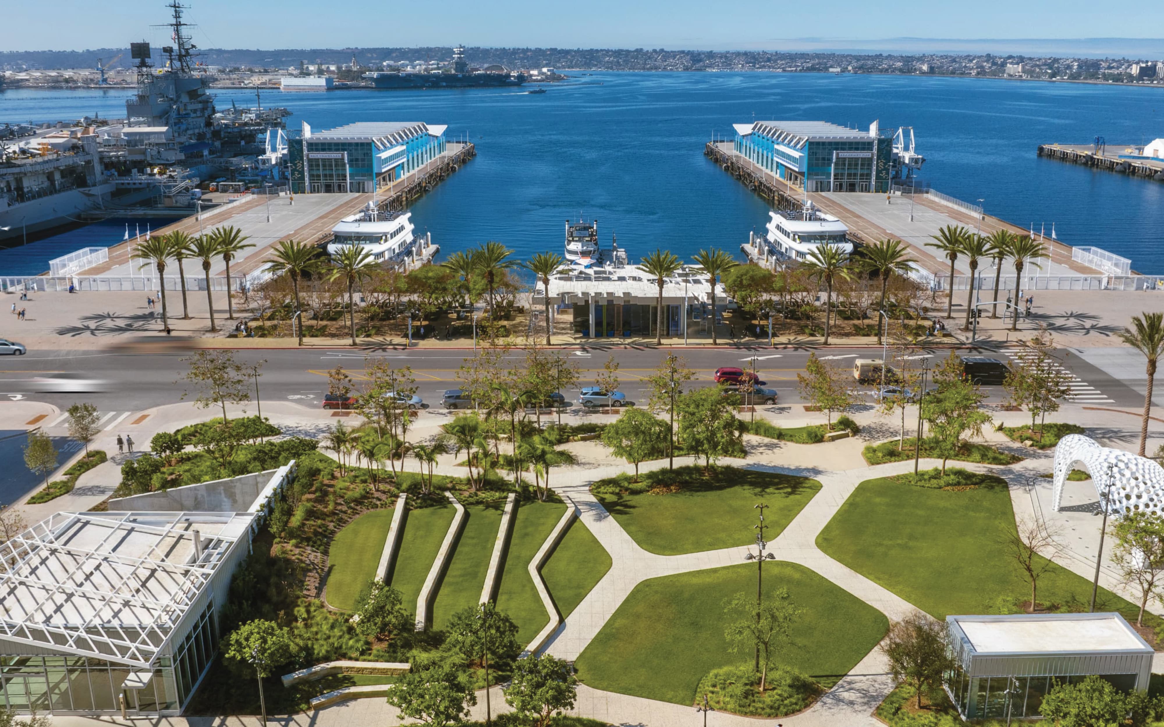 Aerial view of a landscaped waterfront park with terraced lawns and geometric pathways, palm-lined piers with twin blue ferry terminal buildings and docked boats, the USS Midway Museum visible across the harbor, and downtown San Diego on the horizon.