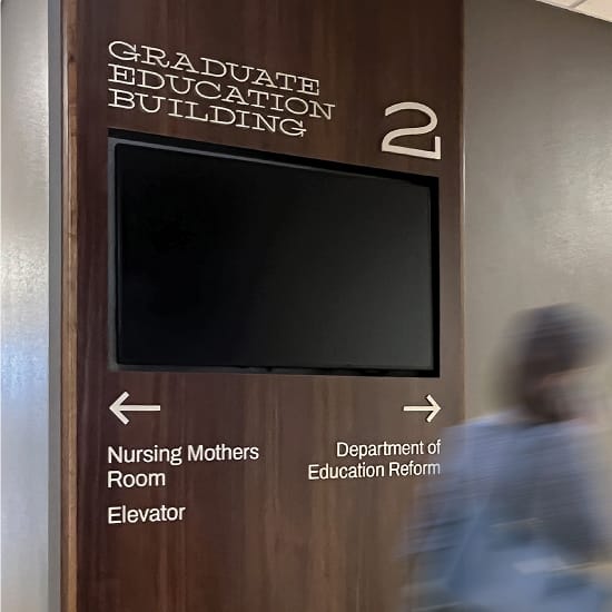 Wayfinding sign on a wood-paneled wall in the Graduate Education Building, Floor 2. A mounted TV screen is centered on the panel. Below it, directional arrows point left to the Nursing Mothers Room and Elevator, and right to the Department of Education Reform.
