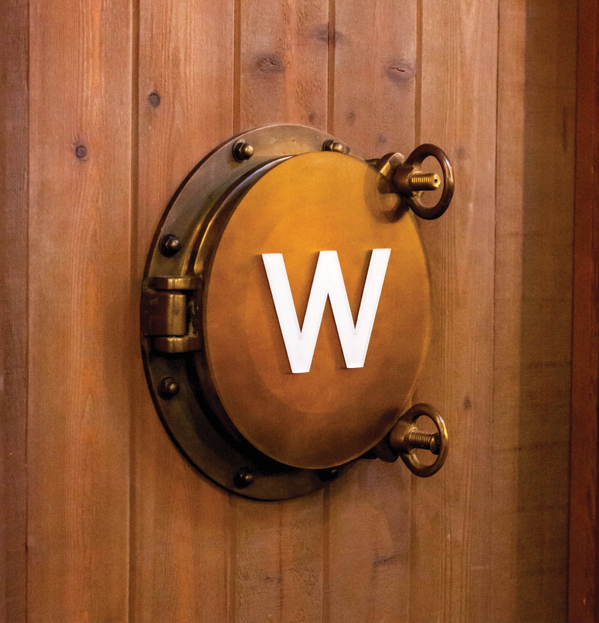 A dimensional white "W" letter mounted on an antique brass porthole fixture affixed to a warm cedar wood wall, serving as a themed restroom identification sign — exemplifying RSM Design's specialty graphics and experiential graphics and branded environments approach to wayfinding and signage design within the food and beverage and mixed-use markets