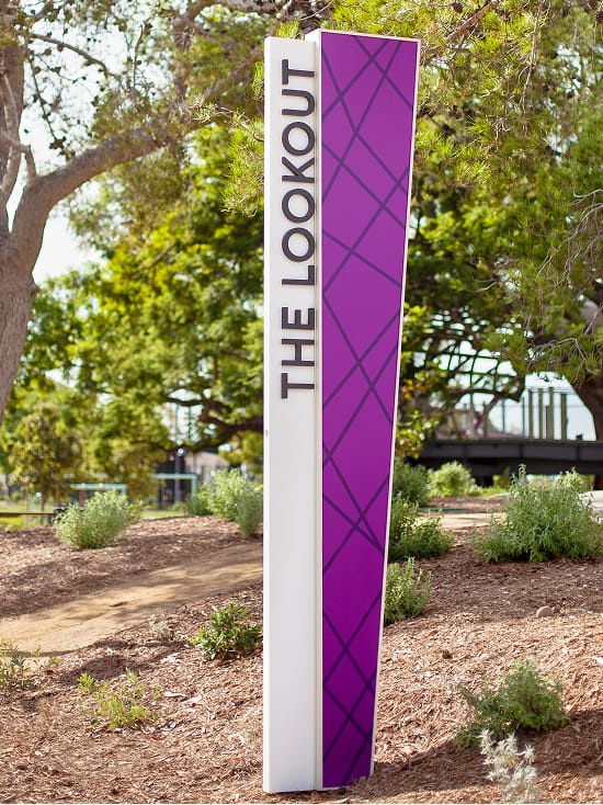 Tall vertical pylon identity sign for The Lookout featuring a bold purple panel with diagonal crosshatch pattern and white dimensional lettering, installed in a park landscape in Irvine, California, designed by RSM Design

