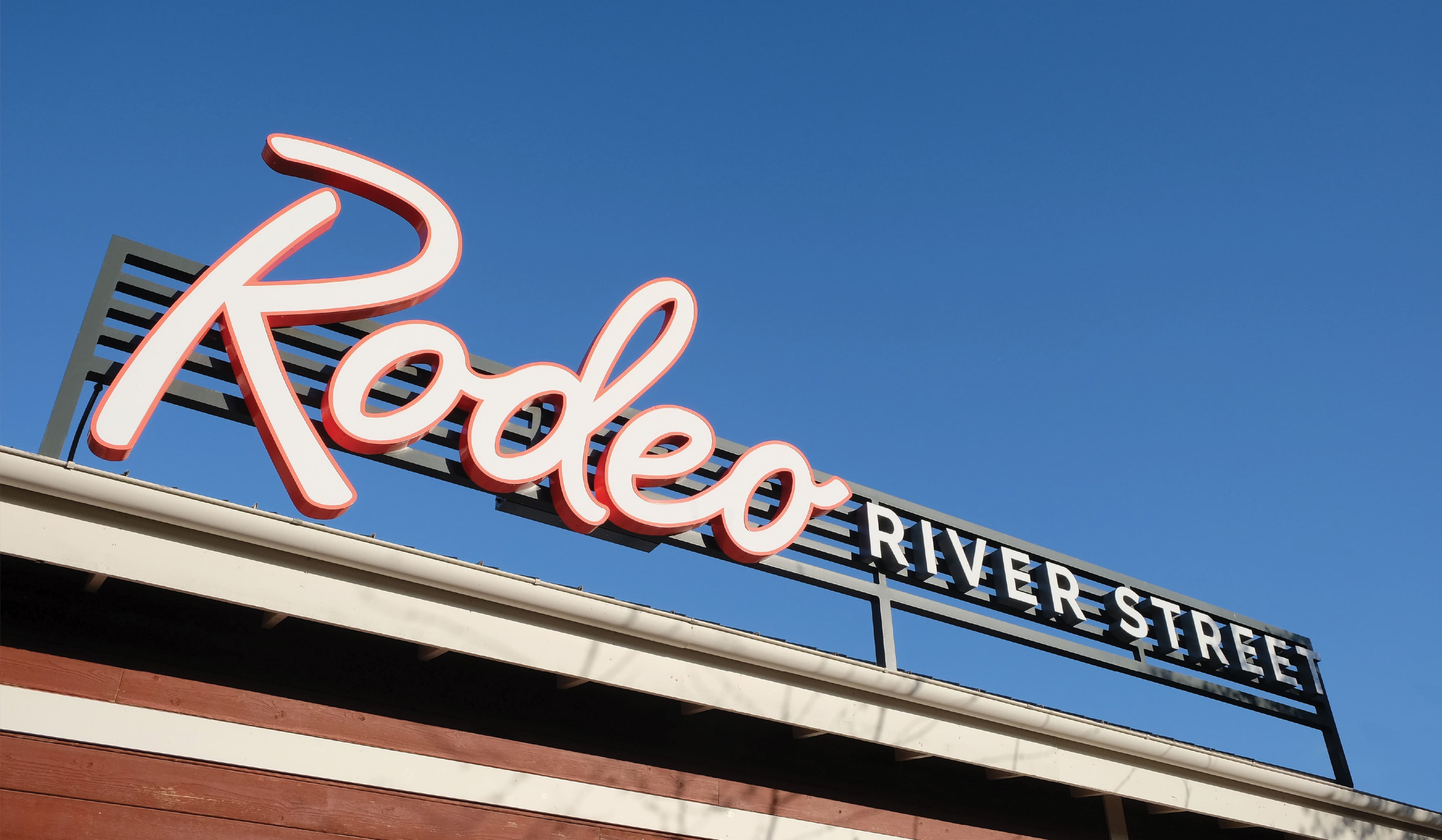 RSM Design large-scale rooftop signage for Rodeo at River Street Marketplace, featuring oversized illuminated dimensional script lettering against a clear blue sky, integrated with a metal address band sign, exemplifying landmark identity signage, experiential graphics, and branded environment design for food and beverage and mixed-use retail placemaking.
