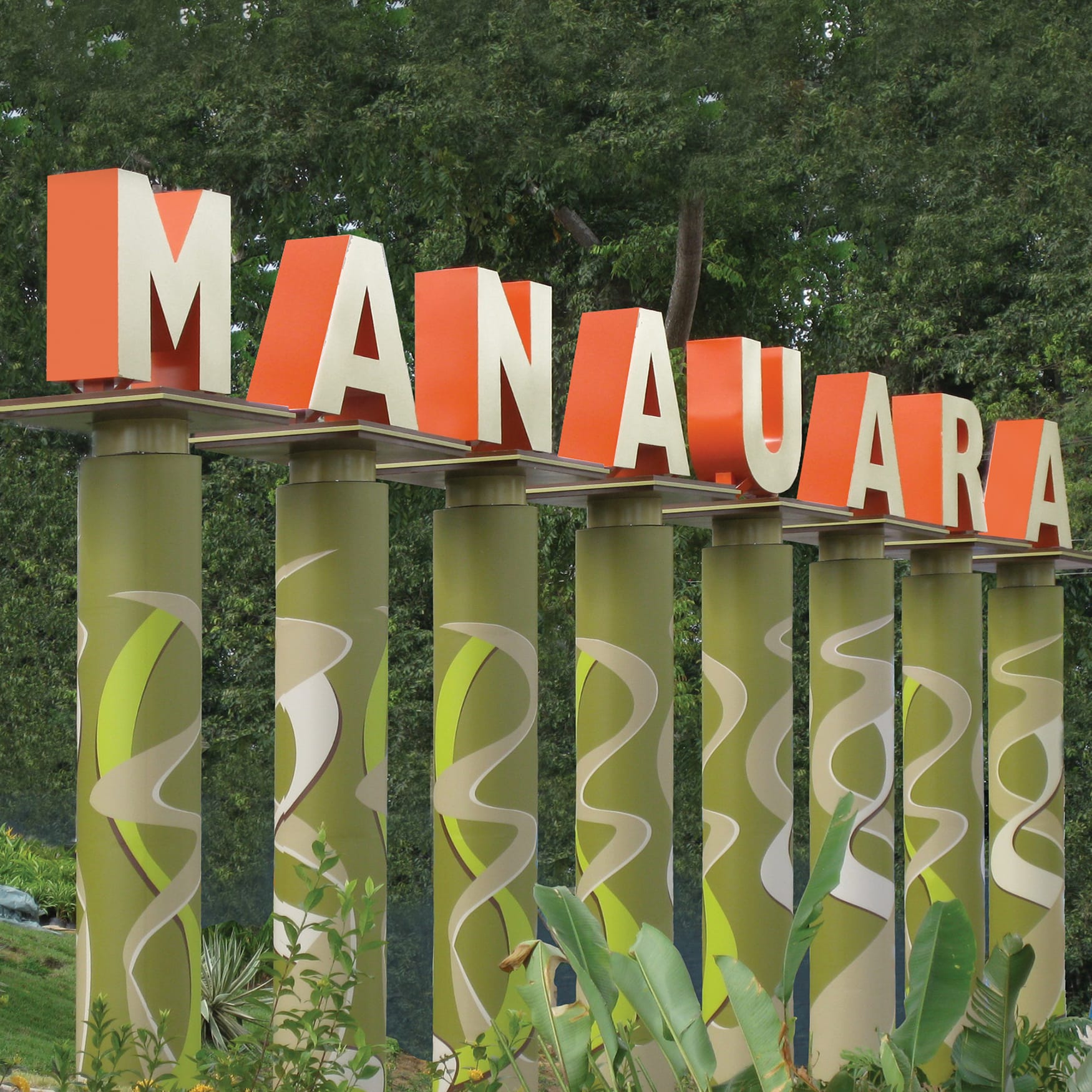 Large outdoor monument sign spelling “MANDAUARA” with bold orange dimensional lettering mounted above sculptural green columns featuring organic wave patterns, surrounded by lush tropical landscaping and trees.